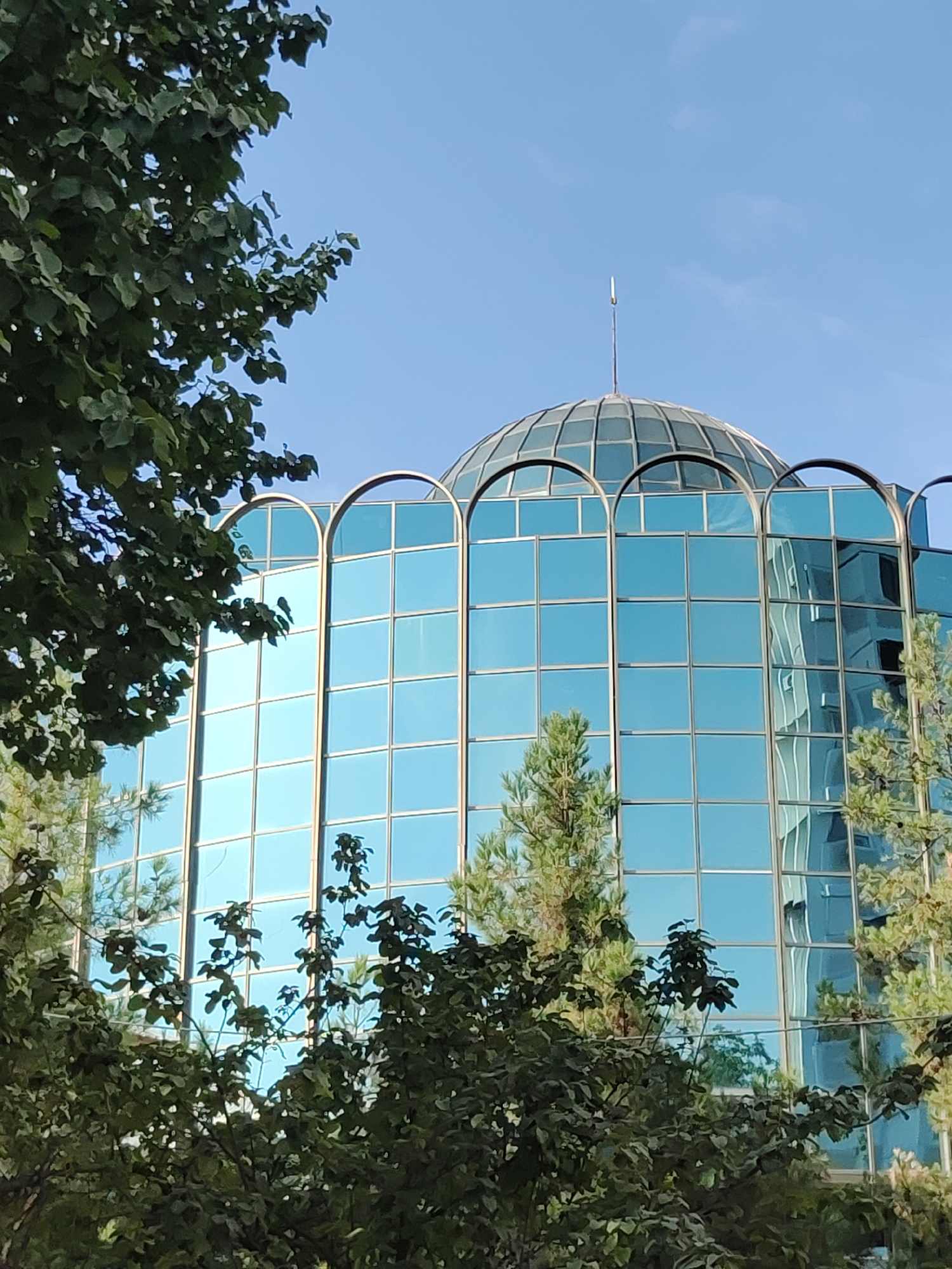 The image shows a modern building with a glass facade and a dome, surrounded by trees and a clear blue sky. The building appears to be made of glass panels reflecting the surrounding environment. A thin, pointed antenna is visible on top of the dome. The building's architecture is contemporary, with arched window frames on the upper floors. The surrounding vegetation is lush and green, suggesting a park or garden area. The scene conveys a sense of urban development within a natural setting. This building is the Presidential Palace of Uzbekistan, located in Tashkent.