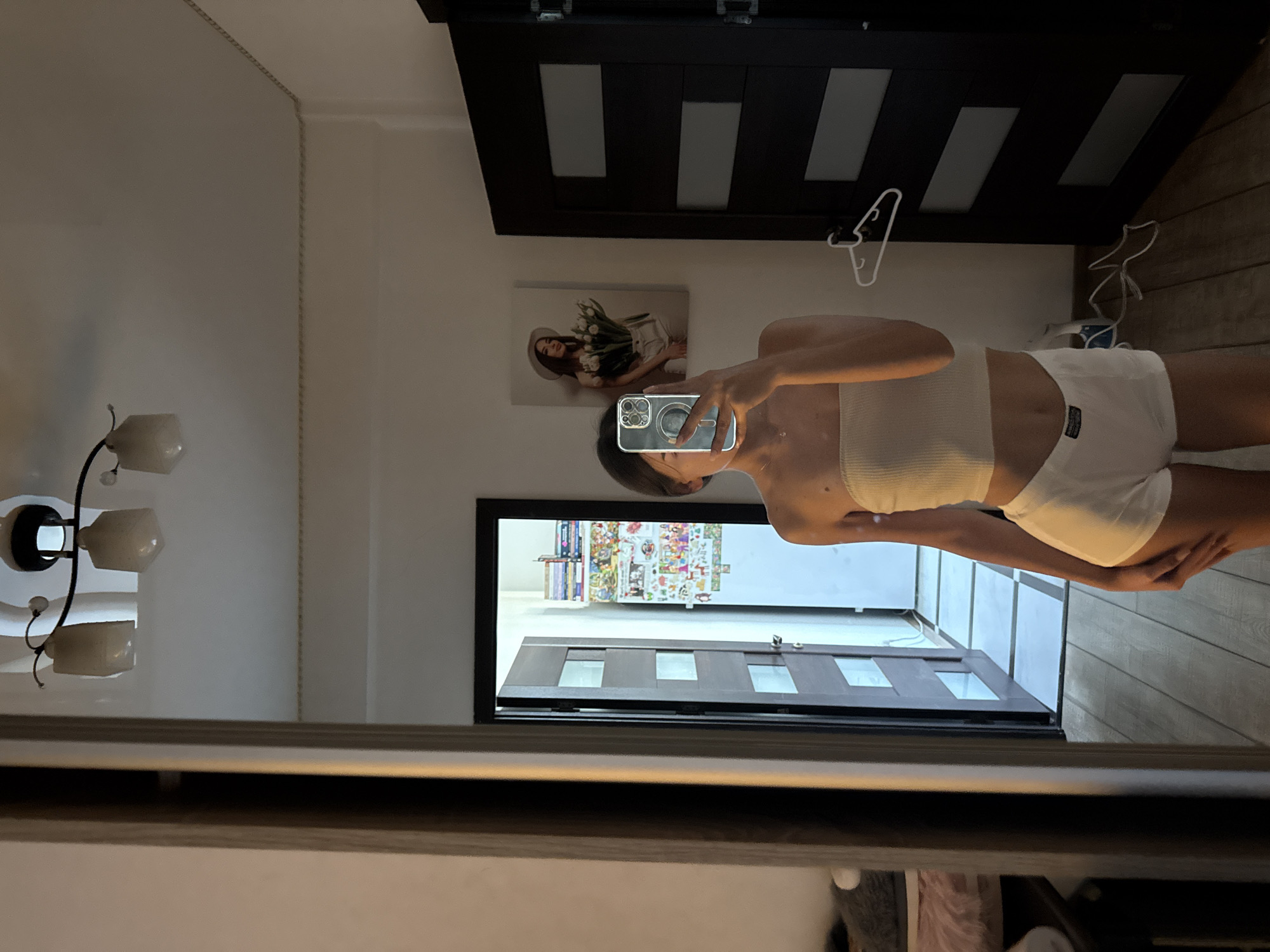 A young woman, visible from the chest down, stands in front of a mirror in what appears to be a residential setting, possibly a bedroom or dressing area. She is wearing a white bandeau top and white shorts. Her reflection shows her holding a smartphone, suggesting she is taking a selfie. In the background, reflected in the mirror, is a doorway leading to another room, a poster on the wall depicting a woman with flowers, and a dark-colored door. The lighting is dim, creating shadows. There is no discernible indication of the city of Chișinău, Moldova, within the image itself.
