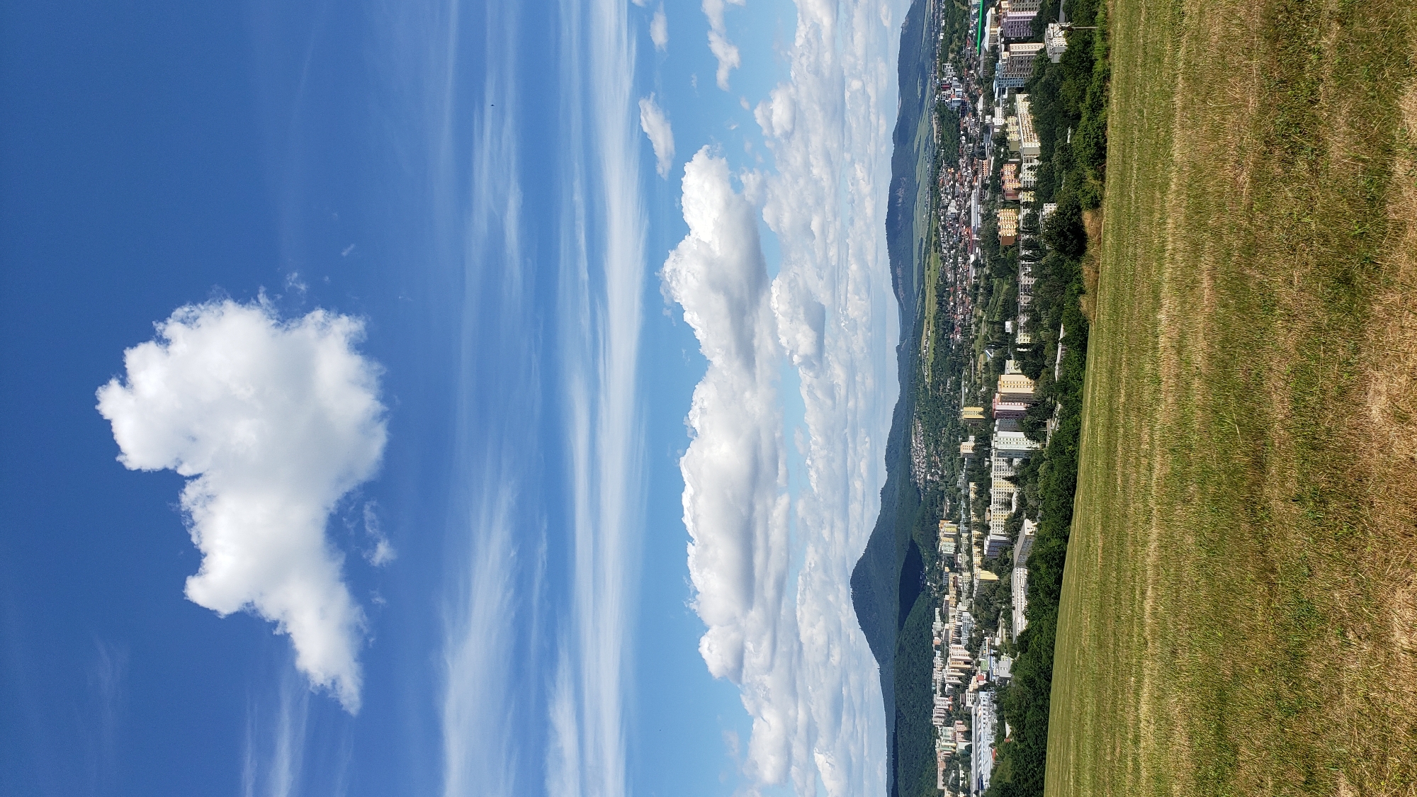 This image is a landscape photograph taken from a grassy hillside overlooking the city of Prešov, Slovakia. The foreground is dominated by a field of dry, yellowing grass, suggesting a summer or late summer season. In the mid-ground, the city of Prešov unfolds with a mix of modern apartment buildings in various colors, interspersed with smaller houses and patches of trees. The city is nestled at the foot of a range of rolling, green hills. The sky above is a vibrant blue, adorned with scattered white cumulus clouds and thinner, wispy cirrus clouds streaking horizontally across the upper portion of the frame. One large, distinct cumulus cloud is prominently positioned in the top center of the image. The overall scene depicts a clear, sunny day with a broad view of urban development against a natural backdrop.