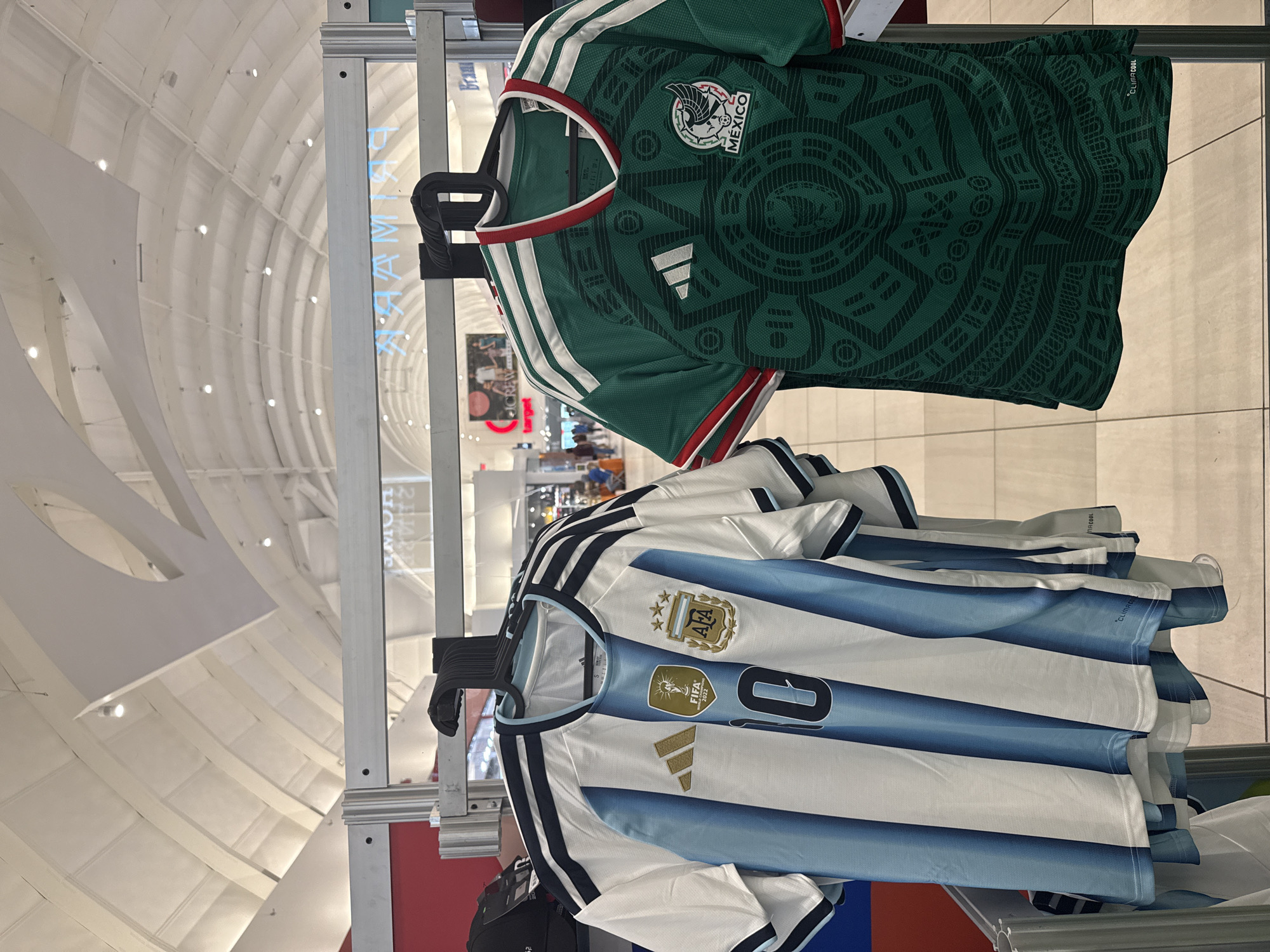 The image displays two national soccer team jerseys, an Argentinian and a Mexican one, hanging on a metal retail display rack within a brightly lit indoor shopping environment. The Argentinian jersey on the left is predominantly white with vertical sky-blue stripes, featuring the Adidas logo, a gold FIFA World Cup 2022 winner's badge, the Argentinian Football Association (AFA) crest with three gold stars, and the number '10' on the front. Black Adidas stripes adorn the shoulders. The Mexican jersey on the right is dark green with an intricate darker green pattern across its body, displaying the white Adidas logo, white Adidas stripes on the shoulders, and the FMF crest with "MÉXICO" below. Both jerseys are suspended on black plastic hangers.
The background reveals a large retail space with a high, vaulted white ceiling illuminated by numerous recessed spotlights. Reflections in a visible glass or shiny surface show reversed text, identifying prominent retailers such as "PRIMARK", "TARGET", and "JCREW," suggesting a major shopping mall. Faint, blurred figures of people can be seen in the distance, indicating other shoppers. The floor consists of light-colored tiles with visible grout lines. Based on the identifiable retailers and the architectural style, the location is characteristic of a large shopping complex like Sawgrass Mills, situated in Sunrise, Florida, United States of America.