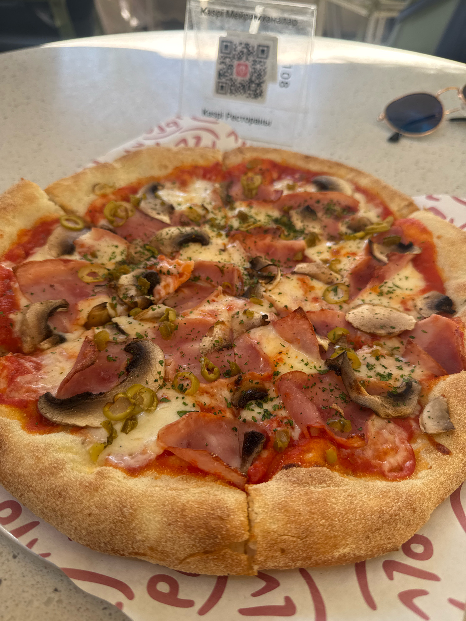 A pizza is displayed on a table in Almaty, Kazakhstan, placed on a paper liner with red lettering. The pizza is topped with sliced mushrooms, ham, green pepper rings, and herbs. In the background, a transparent sign with a QR code and Cyrillic text is visible. A pair of sunglasses is also on the table.
