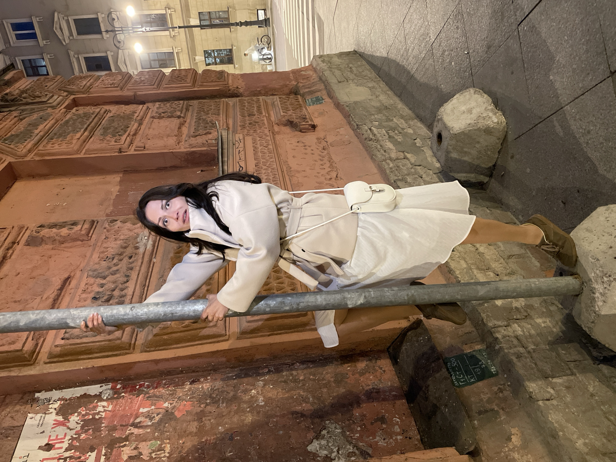 A young woman with long dark hair, wearing a light-colored coat and a white dress, is positioned on a metal pole at an angle, with her legs wrapped around it. Her left hand grips the pole, while her right arm is extended. Her mouth is open in a surprised expression, and her eyes are wide. She is wearing brown shoes and carrying a small white crossbody bag.
The woman is in an urban environment at night. She is standing on a textured sidewalk in front of a building with a reddish-orange, textured facade. The building has decorative elements and windows. To the left of the woman, the building's wall is covered in torn posters. To the right, a street with a crosswalk and a streetlamp are visible, along with the silhouette of a person on a bicycle in the background. The ground consists of large, gray paving stones. The overall scene suggests a candid moment captured on a city street at night, possibly in Saint Petersburg, Russia, given the architectural style and the presence of Cyrillic text on the posters and chalk markings on the wall.