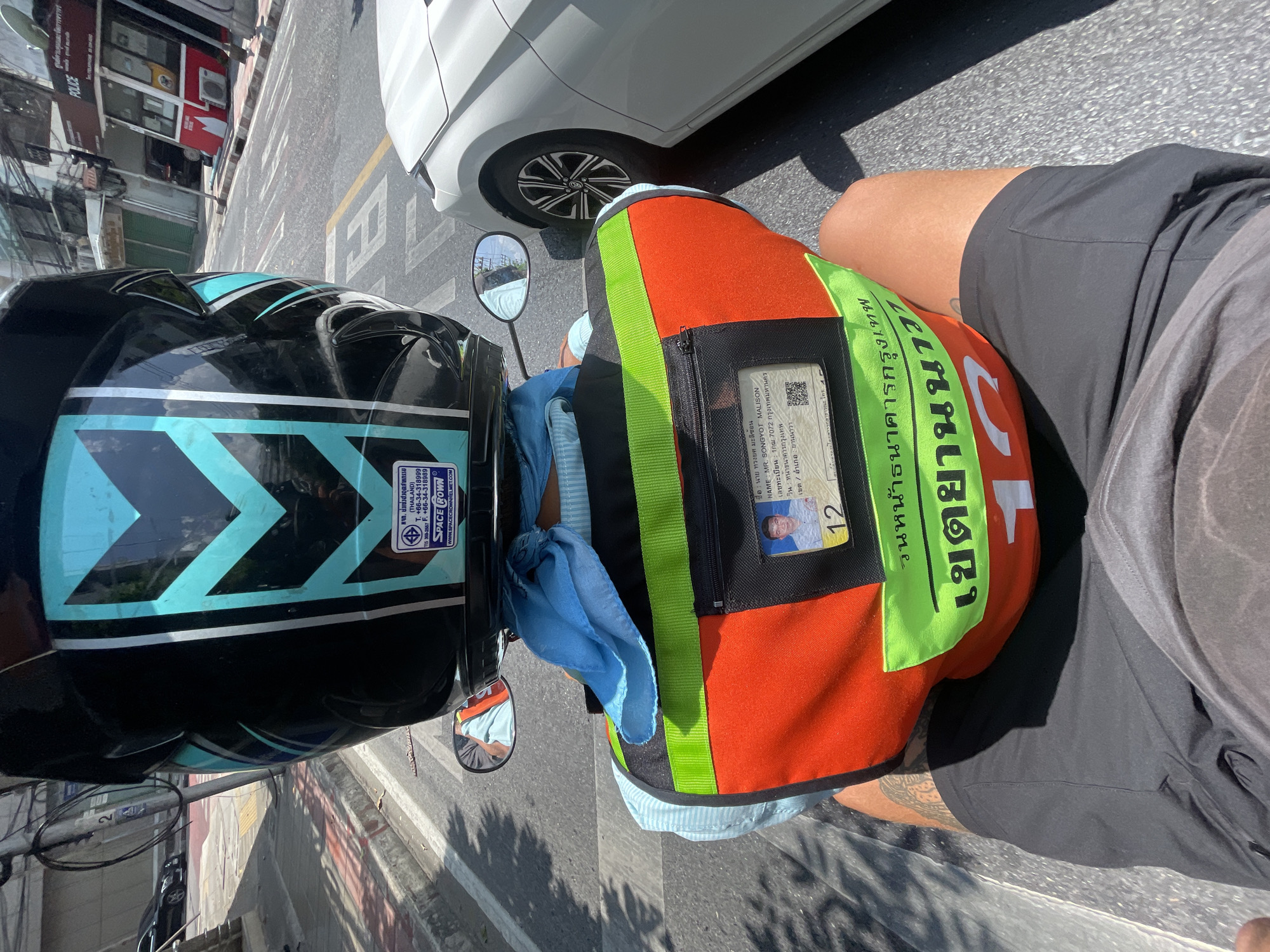 The image depicts a person on a motorcycle in Bangkok, Thailand. The individual is wearing a helmet with blue and black patterns and an orange vest marked with Thai script and the number "12". An identification card is visible in a pocket on the vest, showing a picture of a male and personal details in Thai and English ("NAME: MR. SONGYOT MALISON"). The motorcycle is positioned on a street next to a white car. Street markings are visible on the asphalt. A building with "POLICE" signage in English and Thai is in the background.