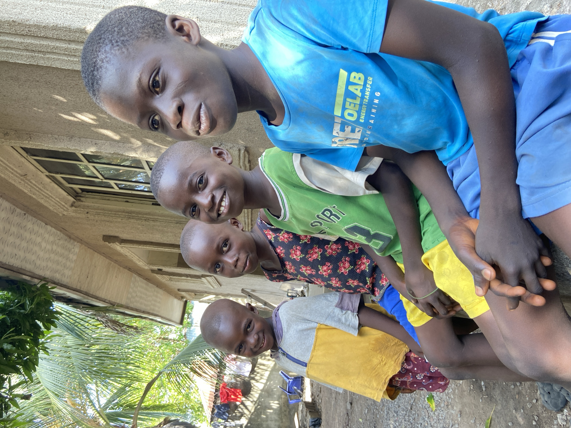 Four Black African boys sit outside a structure in Mararaba, Nigeria, on a bright, sunny day. The boys are stacked vertically in the frame, with the boy at the top wearing a blue T-shirt and shorts, smiling slightly at the camera. Below him, another boy wears a green T-shirt and yellow shorts and has a wide, toothy grin. The third boy, in a floral-patterned shirt, also smiles at the camera. The bottom boy wears a yellow and grey shirt, brown pants, and smiles as well. The boys appear to be sitting closely together. Behind them, a one-story building with a window is visible, and a palm tree extends into the frame from the left. The ground is covered in gravel, and there are some chairs and other objects in the background under a partial canopy.