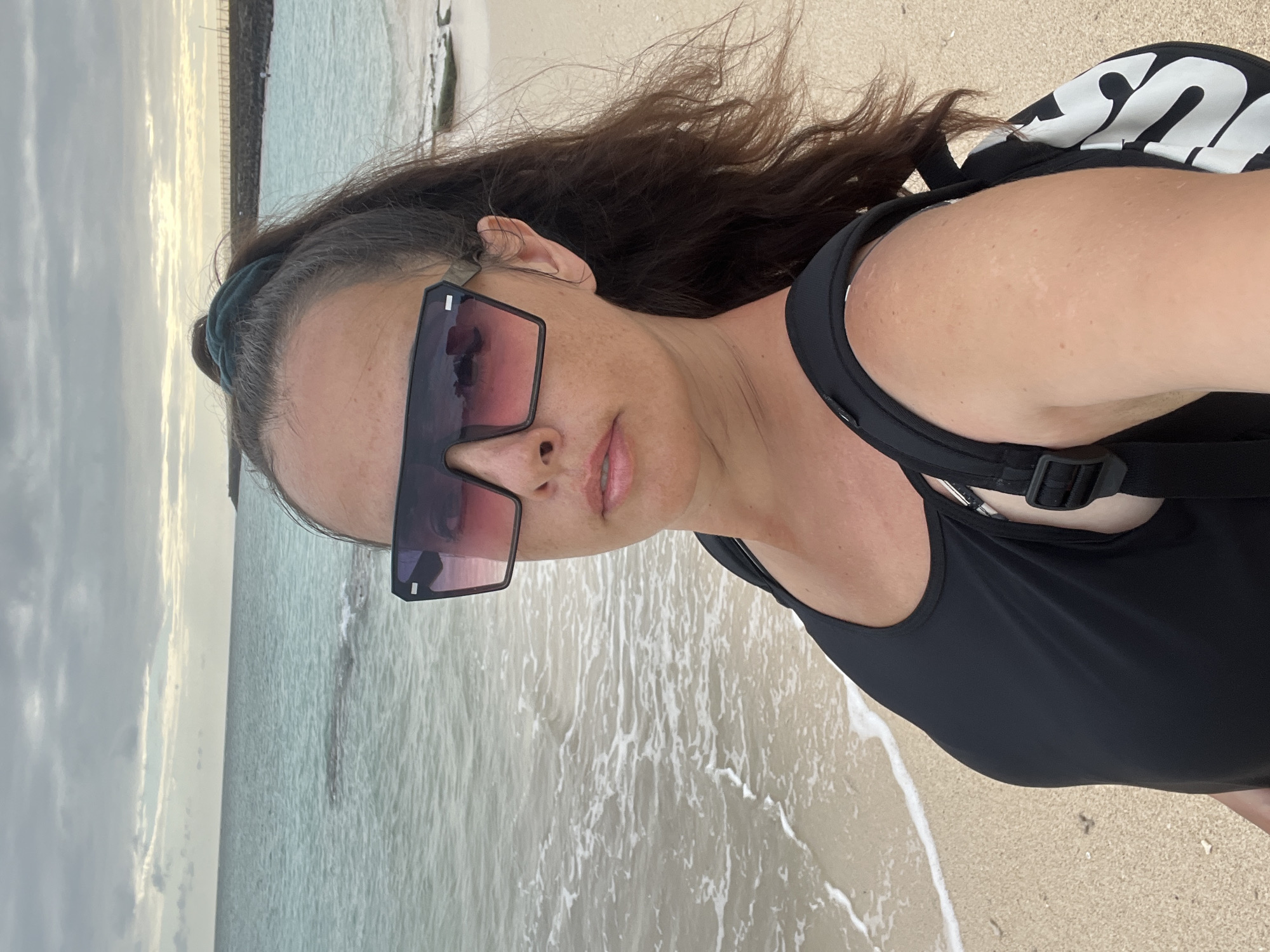 A woman with dark brown hair pulled into a ponytail and wearing large, dark sunglasses stands on a sandy beach, facing the camera. She is wearing a black swimsuit or tank top and a black backpack with white lettering. The ocean with gentle waves is to her left, and the sky is overcast with some sunlight breaking through on the horizon. The location is identified as Kuta, Indonesia.