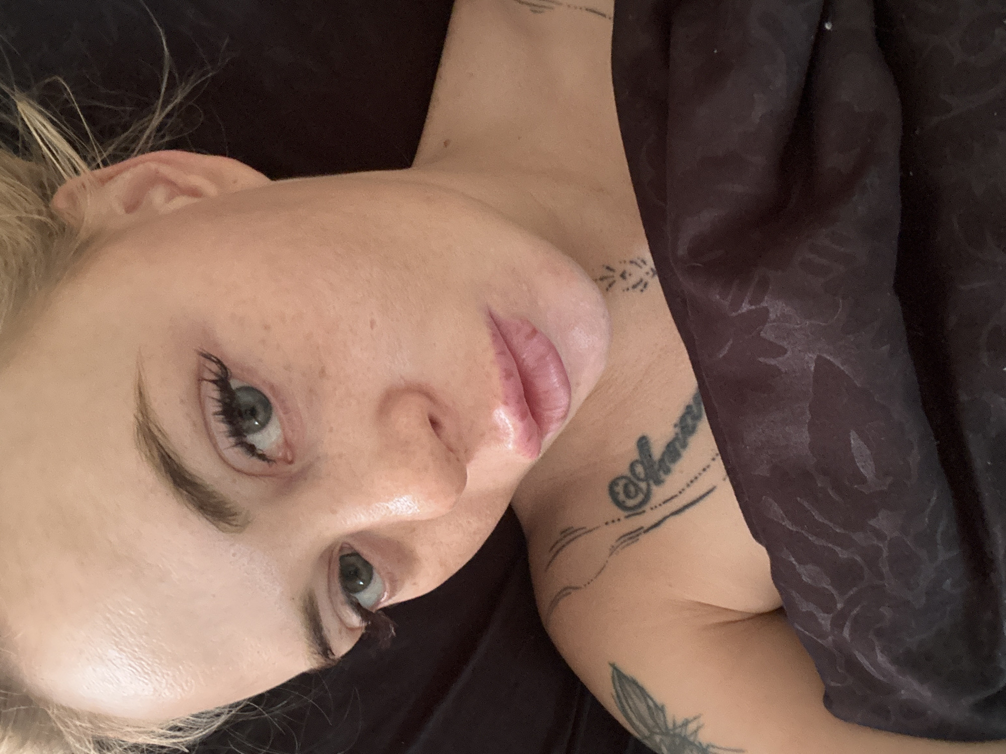 A close-up, high-angle shot shows a woman's face and upper torso, partially covered by a dark, patterned blanket. The woman, with blonde hair pulled back, has light skin with freckles and green eyes. Her lips are slightly parted, revealing a pink hue. She is wearing what appears to be light makeup, including eyeliner and mascara. Visible on her left shoulder and chest are several tattoos. The background is dark, suggesting an indoor setting. There are no other individuals or objects of note in the frame. The lighting is soft, illuminating the woman's face from the front.