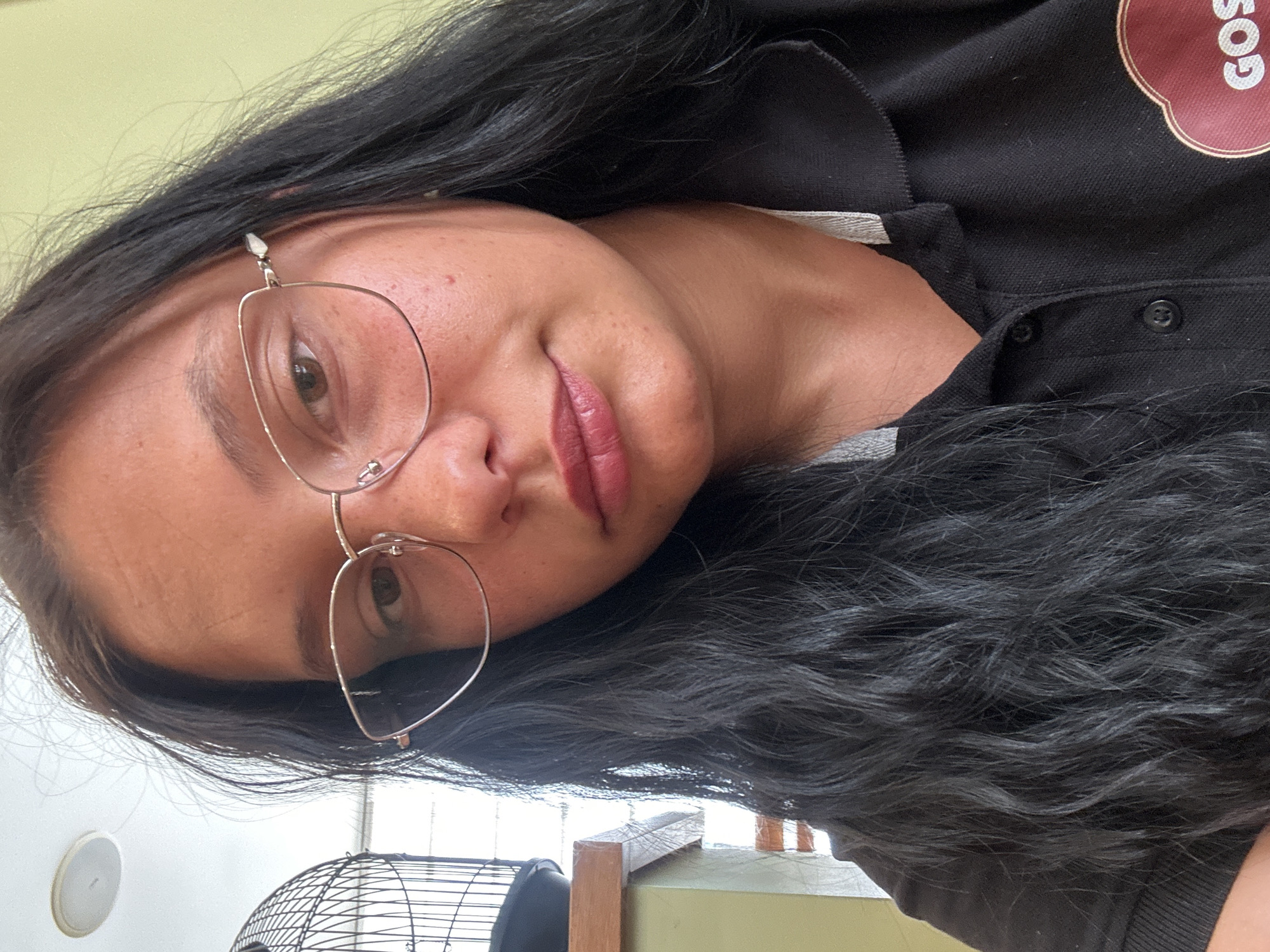 A woman is captured in a close-up, slightly angled shot. She is wearing wire-rimmed glasses and has dark hair. She is wearing a black collared shirt with a logo patch on the upper right side. In the background, a white wall and a fan can be seen. The location cannot be determined to be in Sofia, Bulgaria, based on the visual information in the image.