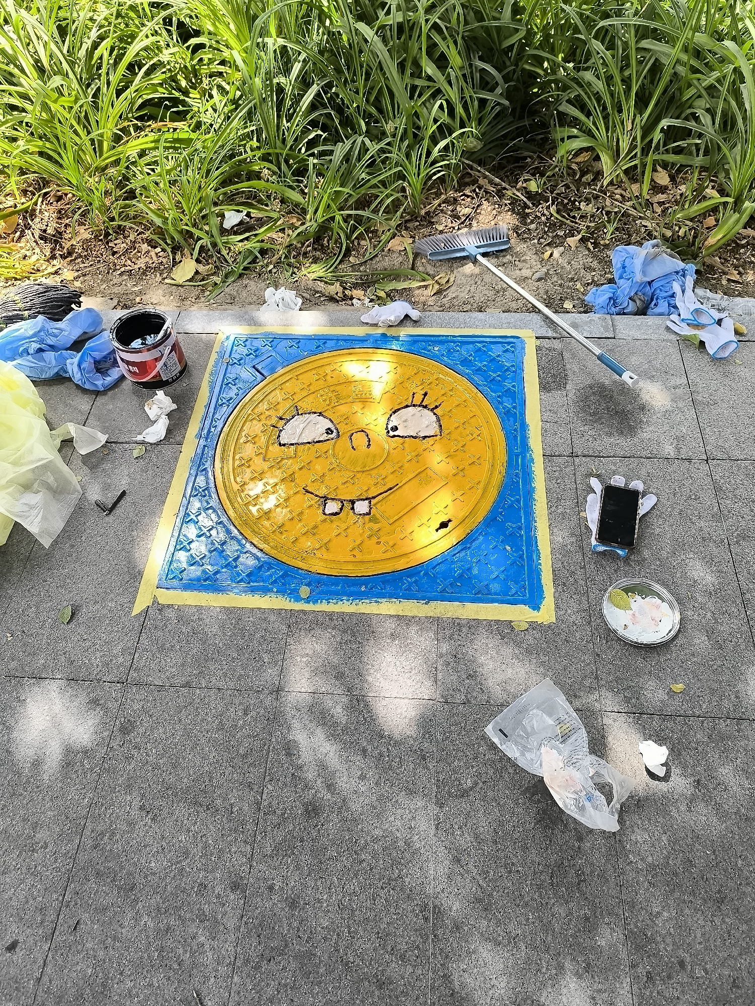 The image depicts a manhole cover on a paved city sidewalk, decorated with a drawing of SpongeBob SquarePants. The manhole cover is painted bright yellow with a blue border, and a SpongeBob face is drawn on it with black lines. Yellow tape surrounds the manhole cover, suggesting it was recently painted or is in the process of being decorated.
Various art supplies and cleaning materials are scattered around the manhole cover. A can of black paint, some crumpled paper towels, and blue plastic bags are visible to the left. A broom with a long handle and a blue brush head is positioned to the right of the manhole cover, with more blue plastic bags and white gloves nearby. Further to the right, a smartphone is placed on top of a white object, and a small dish with paint remnants and a pink flower-like design is situated on the ground. A crumpled transparent plastic bag and some scattered white material are also present.
The background shows a strip of lush green grass and foliage, typical of an urban green space or park area. The sidewalk is made of grey granite tiles. The scene appears to be outdoors, under natural lighting, indicated by the shadows cast on the ground. The overall impression is that of an impromptu art project or street art activity.
There are no visible people in the image. The location is identified as Ürümqi, China.