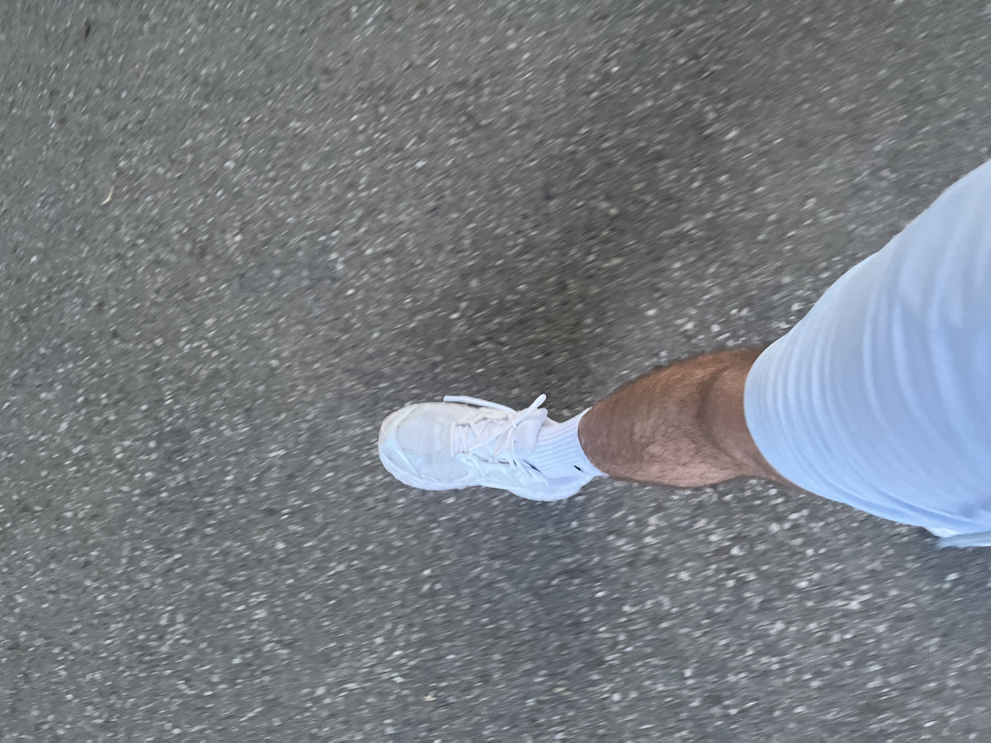 The image depicts a person's foot in a white sneaker and sock on gray asphalt. The person is also wearing white shorts. The specific location in Blagoevgrad, Bulgaria, cannot be determined from the given visual information. The person appears to be in motion.