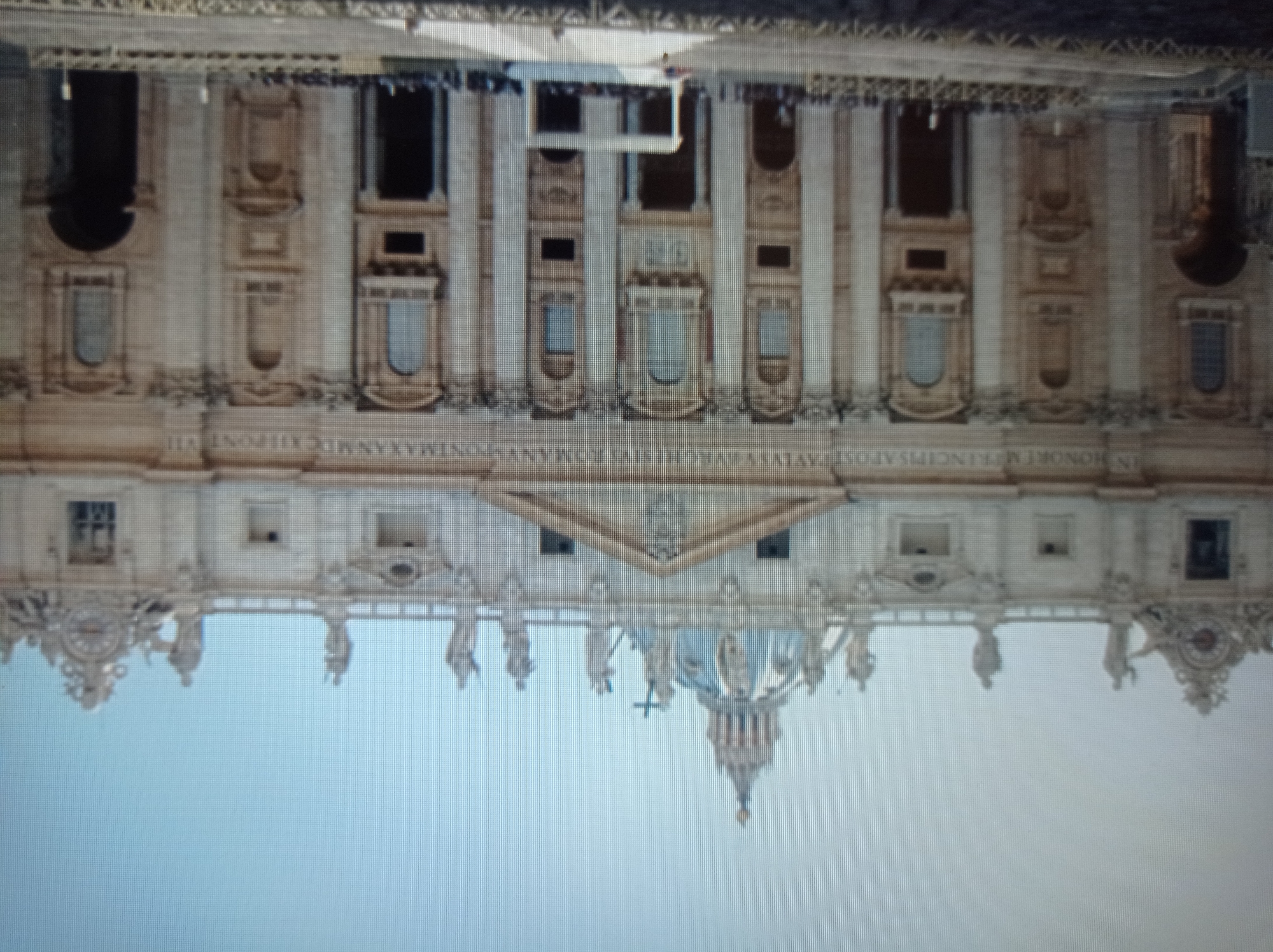 The image shows a wide-angle view of the facade of St. Peter's Basilica in Vatican City, Rome, Italy, under a clear blue sky. The basilica's grand architectural features, including its large dome, numerous statues, columns, and arched entrances, are visible. In front of the basilica, a crowd of people is gathered, facing the building, suggesting a public event or ceremony is taking place. There is a white stage-like structure set up in the plaza. The overall scene conveys a sense of scale and importance, typical of a major religious and tourist landmark. The image does not depict the city of Mukachevo, Ukraine.