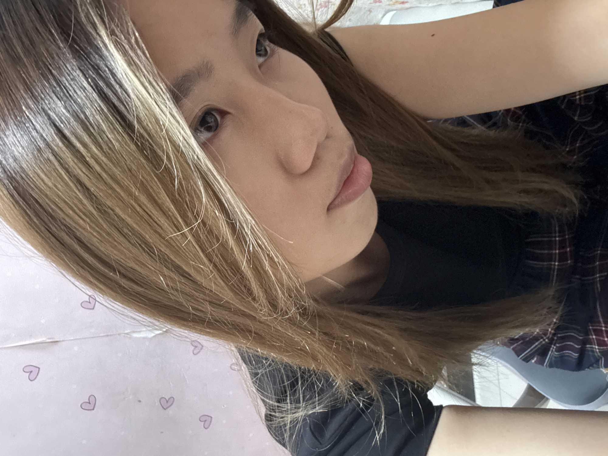 The image shows a close-up of a young Asian woman with long, blonde-brown hair, wearing a black t-shirt. She is looking down and to her right, with her head tilted. The background appears to be a wall with pink wallpaper featuring small, light purple heart patterns. The setting is indoors, possibly a room. There is no indication of the specific location within Shapingba District, China.