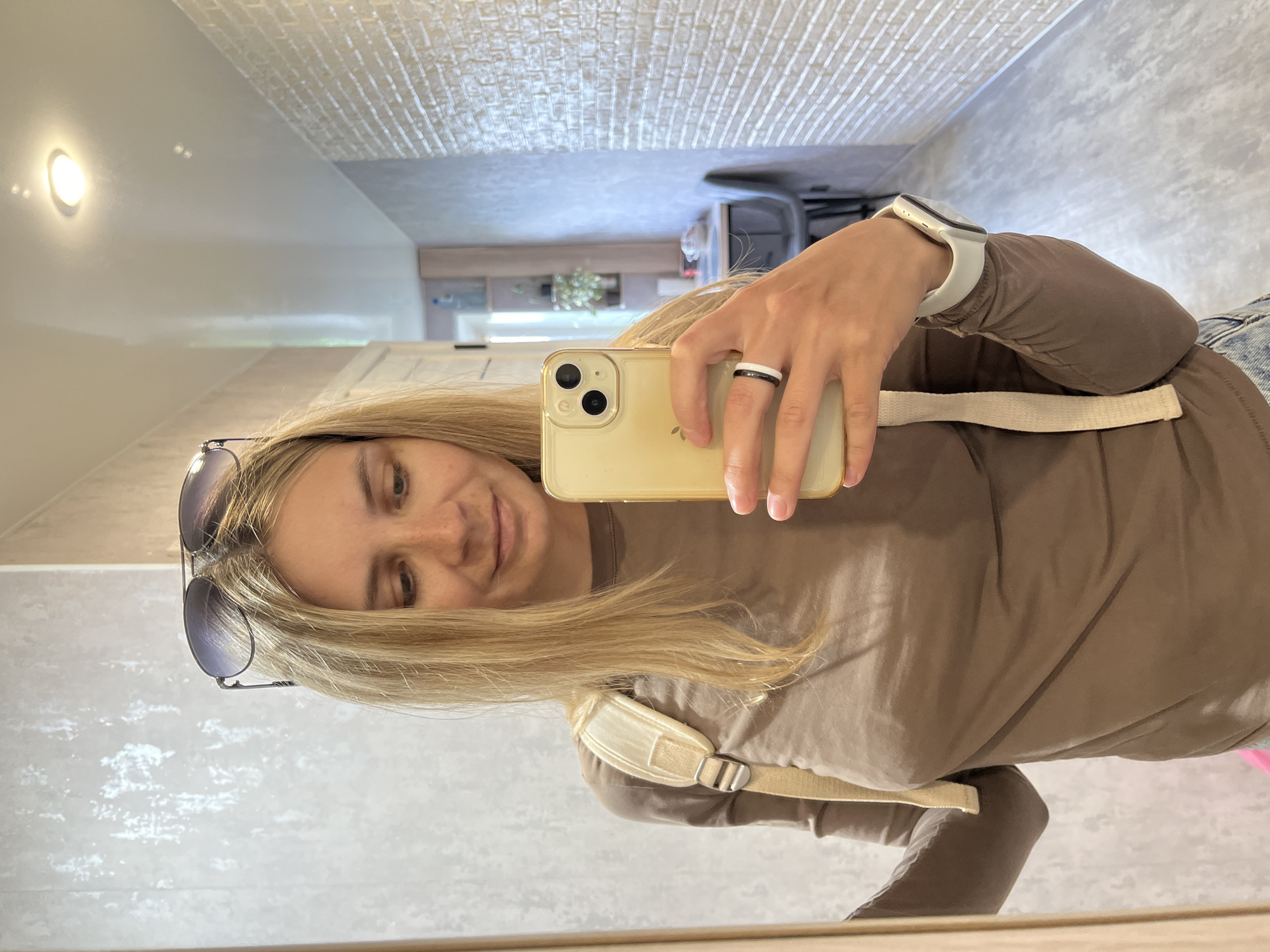 A woman with long blonde hair is captured in a mirror selfie, holding a light-colored smartphone with dual cameras in her right hand. She has a subtle smile and her eyes are directed towards the phone. She is wearing a brown long-sleeved top and jeans, with a beige strap of a backpack or bag visible over her right shoulder. On her left wrist, she wears a white smartwatch, and a black and white ring adorns a finger on her right hand. Sunglasses are perched on top of her head. The reflection shows an indoor setting, likely a hallway or room, with a light fixture on a plain wall and a textured, possibly brick-patterned, ceiling. The background further away in the reflection suggests other interior elements. The scene takes place indoors in Chaikovsky, Russia. No visible text is present in the image.