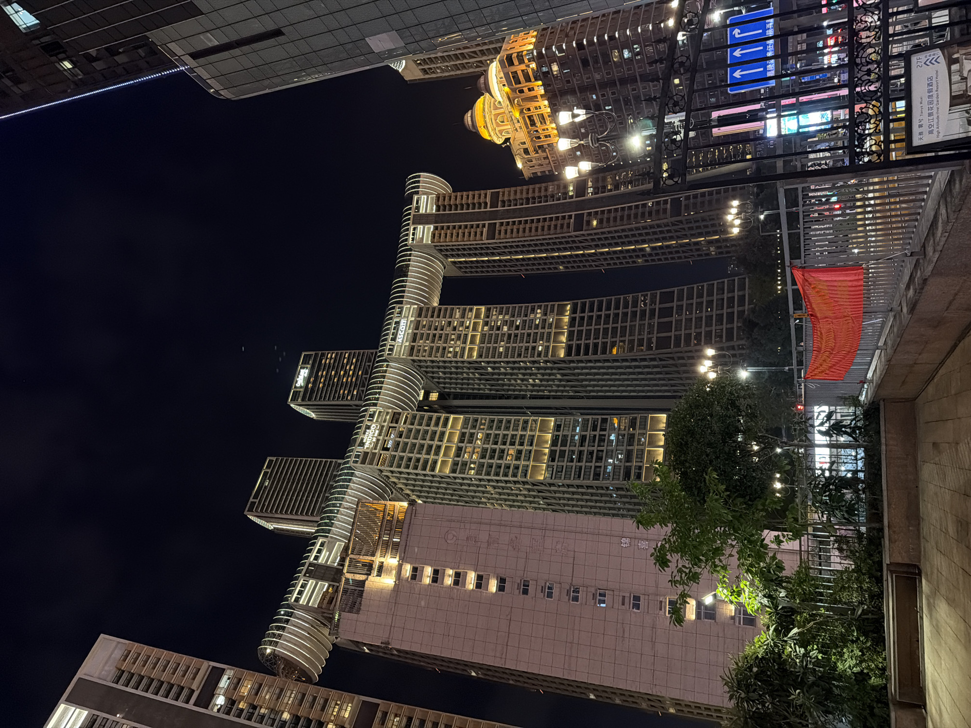 A nighttime exterior view captures the iconic Raffles City Chongqing complex in Yuzhong District, China. The photograph is taken from a slightly low angle, emphasizing the grandeur of the architecture. The central feature is the distinctive "Crystal" structure, a horizontal glass tube connecting four skyscrapers, illuminated with white light. The skyscrapers themselves are tall, modern buildings with numerous windows, many of which are lit, suggesting occupancy.
To the left of the Raffles City complex, a pinkish-beige skyscraper is visible, featuring rows of small, uniformly spaced windows. Faint Chinese characters are discernible on its facade. A green tree with lush foliage stands in the foreground, partially obscuring the lower section of this building.
To the right of the central structure, another skyscraper is visible, predominantly glass and metal, with a grid-like pattern. Further to the right, a building with a golden, domed roof stands out, brightly lit with orange and yellow lights, creating a contrasting architectural style.
In the foreground, a black wrought-iron fence with decorative elements separates the viewer from a lower level. Behind the fence, a red banner hangs, displaying some form of text or pattern. Streetlights and illuminated signs are visible in the background, contributing to the vibrant nighttime urban landscape. A blue directional sign with white arrows is also present, indicating directions. The ground in the immediate foreground is paved with light-colored tiles. The overall scene conveys a bustling, modern cityscape at night.
