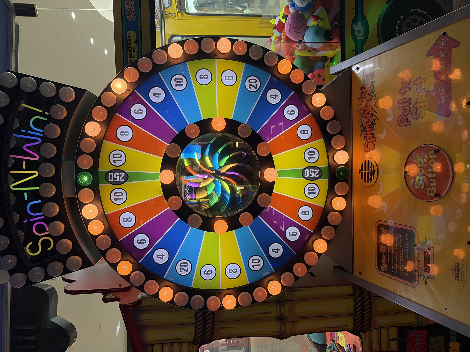 The image shows a "Spin-N-Win" arcade game. The game features a large, illuminated wheel with numbered sections (4, 6, 8, 10, 20, 250). A sign reads "Spin-N-Win! Pull to Start" and has a "Stop Button." A digital display shows "0" tickets. The game's instructions include inserting coins/tokens, pulling the handle to start, pressing the button to stop the lights, and collecting tickets. Other arcade games are visible. The location is not discernible from the image.
