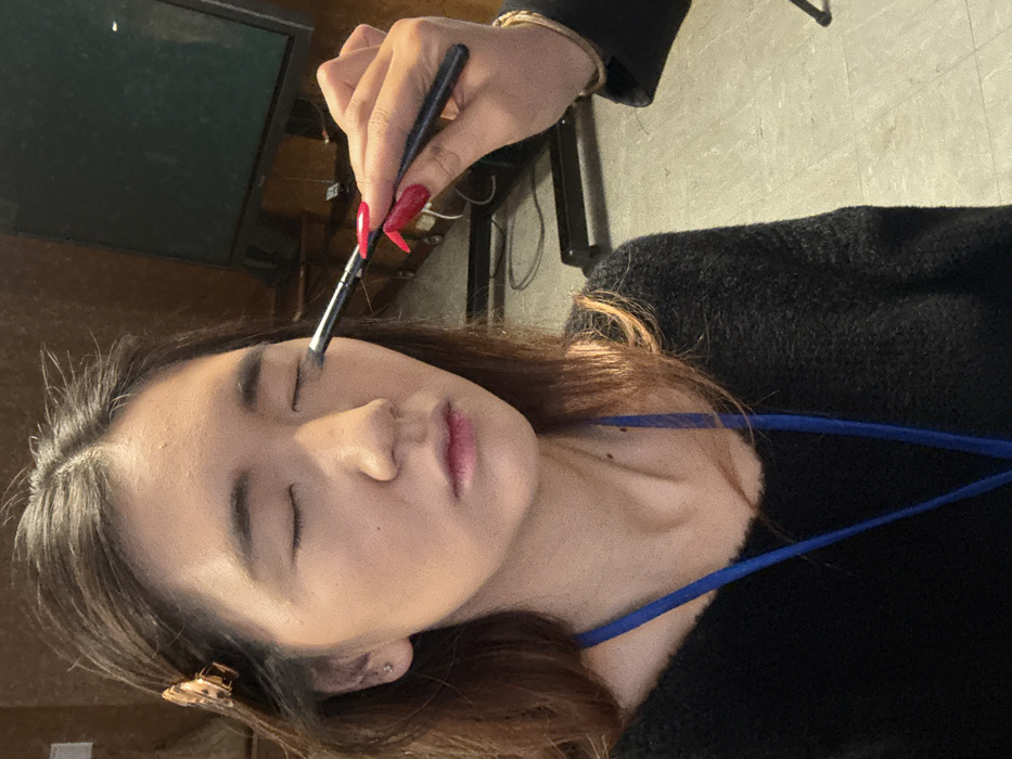 A woman with her eyes closed has makeup applied to her brow with a brush held by a hand with red nails. The woman is wearing a black sweater and a blue lanyard. She has long brown hair with a clip on the side. A television is visible in the background. The hand applying the makeup is also wearing a gold bracelet.