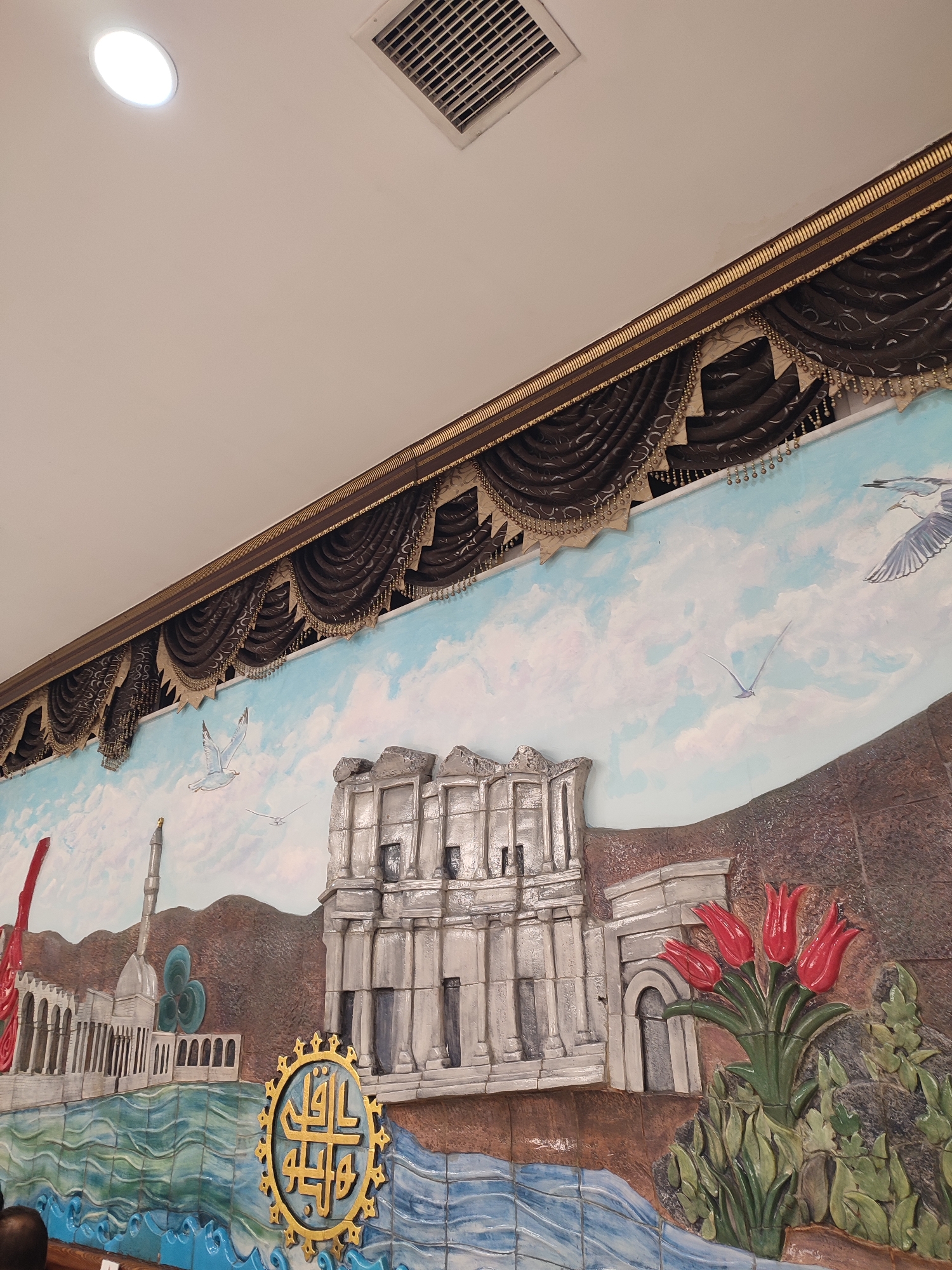 The image depicts a decorative mural on a wall, likely in Istanbul, Turkey, given the architectural style and motifs. The mural features a landscape with a body of water in the foreground, possibly representing the Bosphorus Strait or the Golden Horn. On the left side of the mural, a white building with arches and columns is depicted, resembling a mosque or a historical palace. Adjacent to it is a tall, slender minaret. In the center of the mural is a representation of ancient ruins, possibly a Roman or Byzantine structure, with columns and arched doorways. To the right of the ruins, a cluster of red tulips is rendered with detail, set against a backdrop of textured brown hills. Several seagulls are painted in the sky, which is a light blue with white clouds. A circular golden emblem with Arabic calligraphy is prominently displayed on the water's surface, near the ancient ruins. Above the mural, a section of the ceiling is visible, with a round light fixture and an air vent. Dark brown, ornate curtains with gold trim hang above the mural, suggesting it might be part of a restaurant, hotel, or public space. A small portion of a person's head and shoulder is visible in the lower left corner, out of focus.