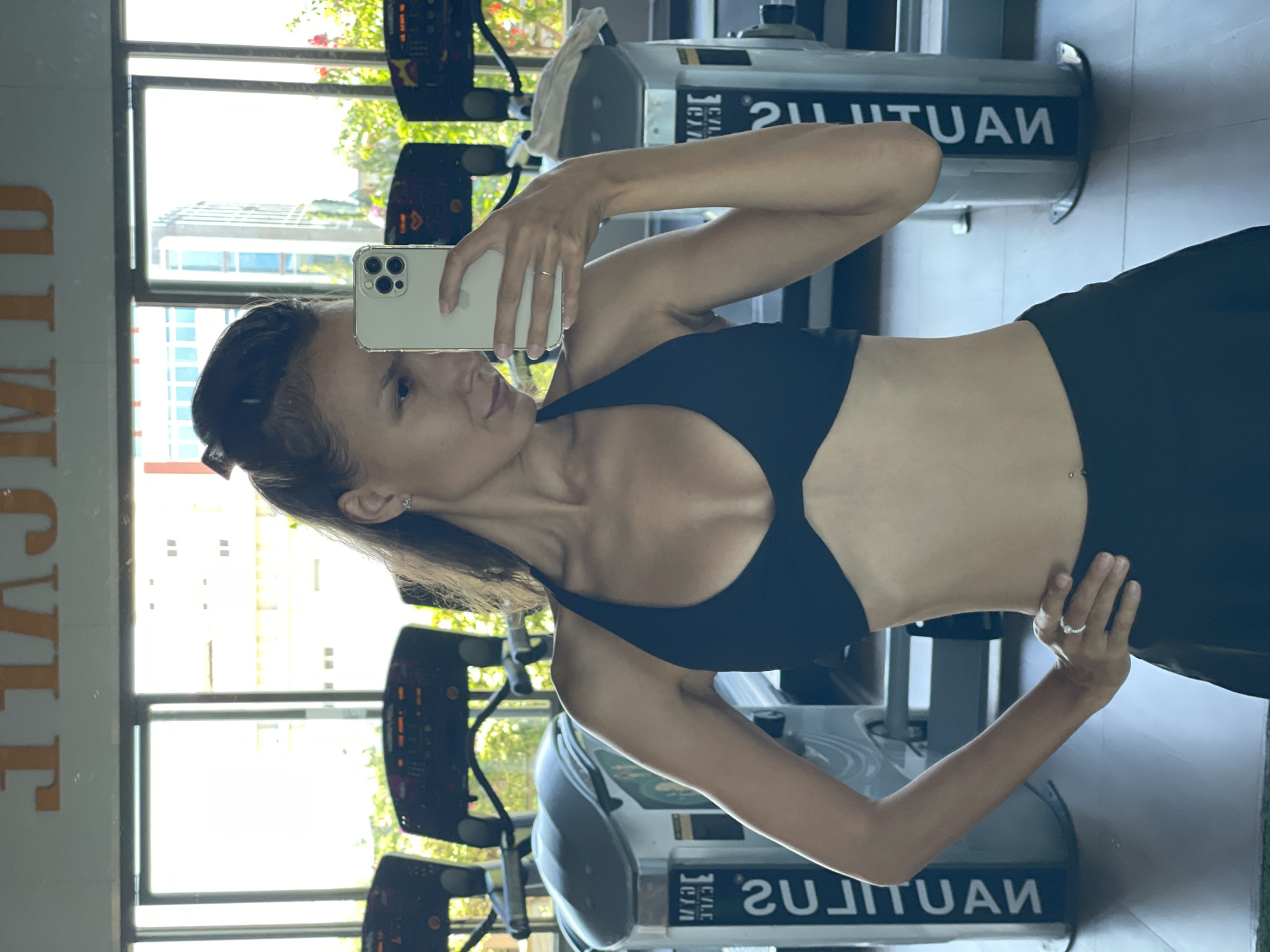 A woman is taking a mirror selfie in a gym. She is wearing a black sports bra and black leggings. She has her hair pulled back in a ponytail and is wearing earrings and rings. In the background, there are treadmills and windows that overlook a city. The gym appears to be located in Ham Thang Ward, Vietnam, as indicated by the text on the treadmills which reads "GYM CAFE" and the architecture visible through the windows.