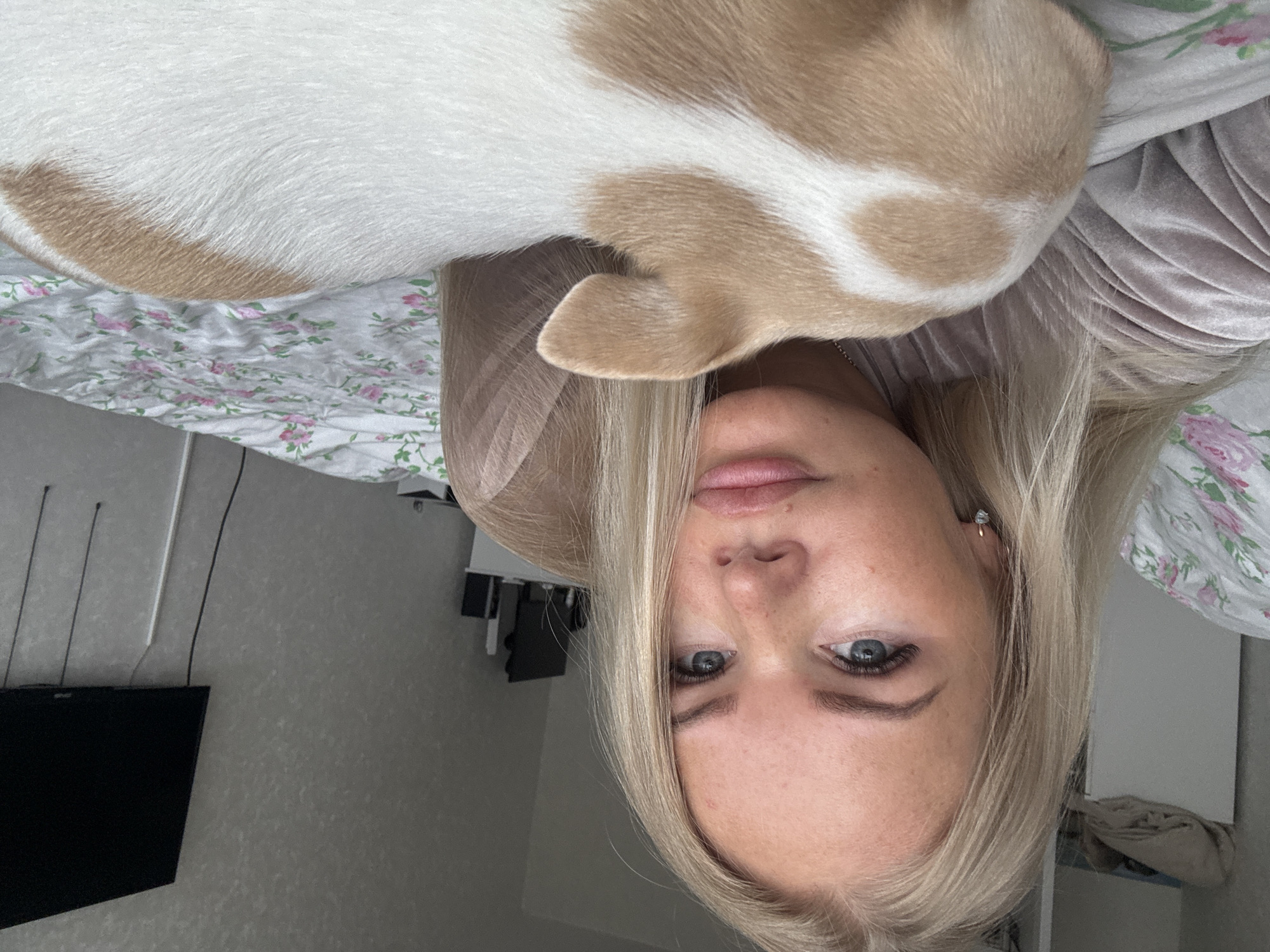 A woman with blonde hair and blue eyes is lying on a bed with a dog. The dog is white and tan, and is lying on the woman's chest. The bed has a floral patterned bedspread. A television is visible on the wall in the background. The location is Unknown, Unknown.
