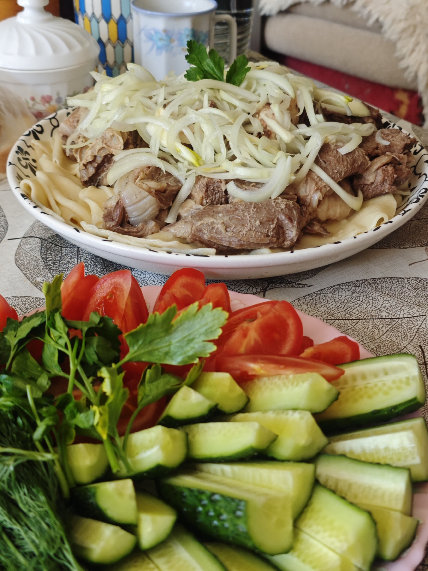 A close-up shot presents a spread of food in what appears to be a home setting. A large, round plate features a dish of what seems to be meat chunks on top of noodles, garnished with slivered onions and a sprig of parsley. Below this plate, a smaller, pink plate holds sliced tomatoes, cucumbers, and fresh green herbs, including dill and parsley. The plates rest on a table with a leaf-patterned tablecloth. In the blurred background, there are kitchen items, including a white covered dish and a couple of mugs. The presence of a woolly object in the top right corner suggests a domestic environment. Given the context of Chaikovsky, Russia, as specified in the request, it is implied that the food may be a traditional dish of the region, such as Beshbarmak.