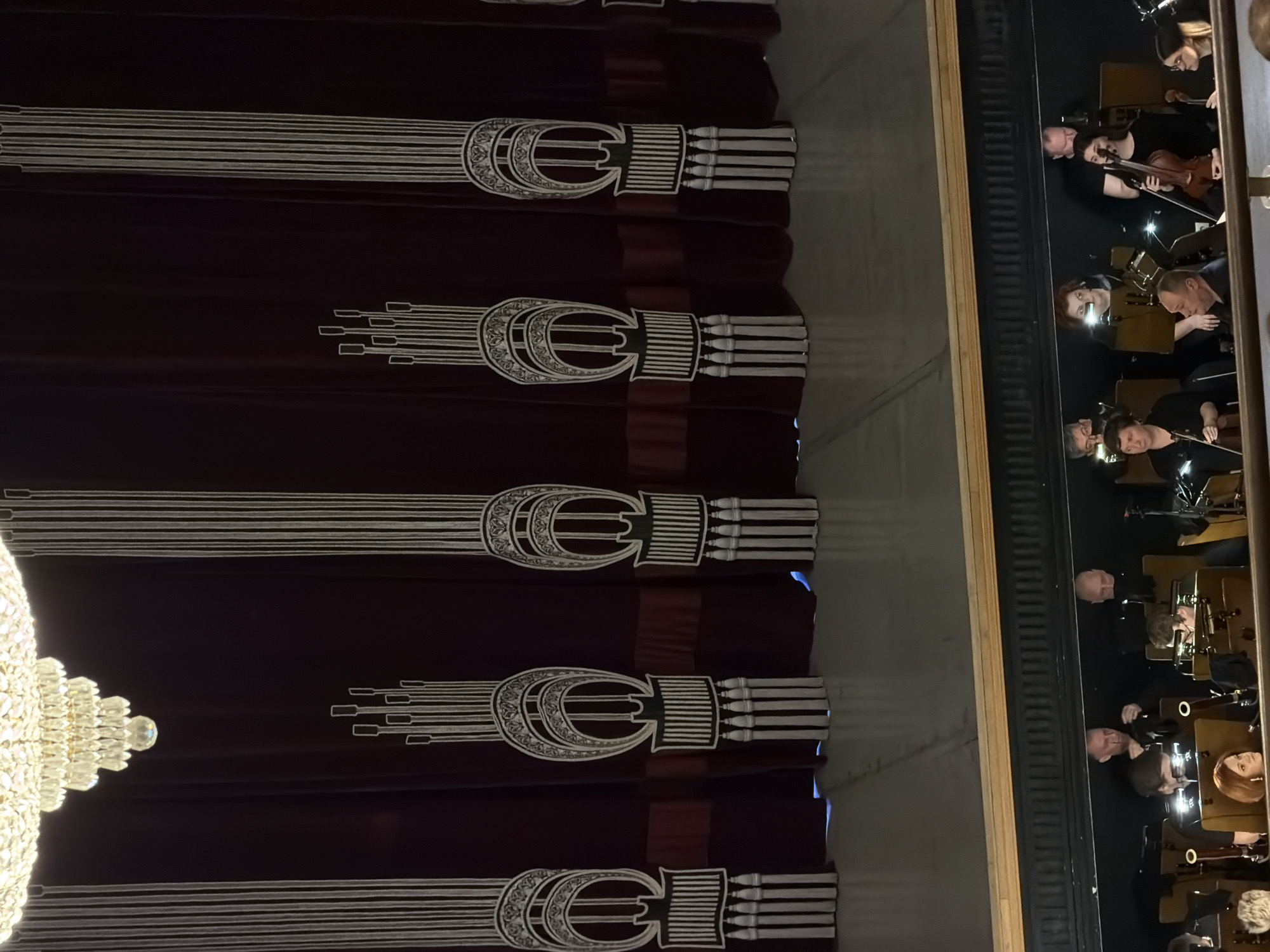 The image depicts the interior of a theatre or performance hall. Dominating the left side of the frame is a large, ornate chandelier. Dark maroon curtains with a recurring silver and white emblem (possibly a stylized double "C" or "E") hang behind the chandelier.
On the right side of the frame, a reflective surface, possibly a mirror or a window, shows a partial view of an orchestra pit. Several musicians are visible, primarily playing violins. They are dressed in black. The positioning suggests they are preparing for or engaged in a performance. The scene is possibly located in the State Opera and Ballet Theatre of the Udmurt Republic (Izhevsk, Russia).