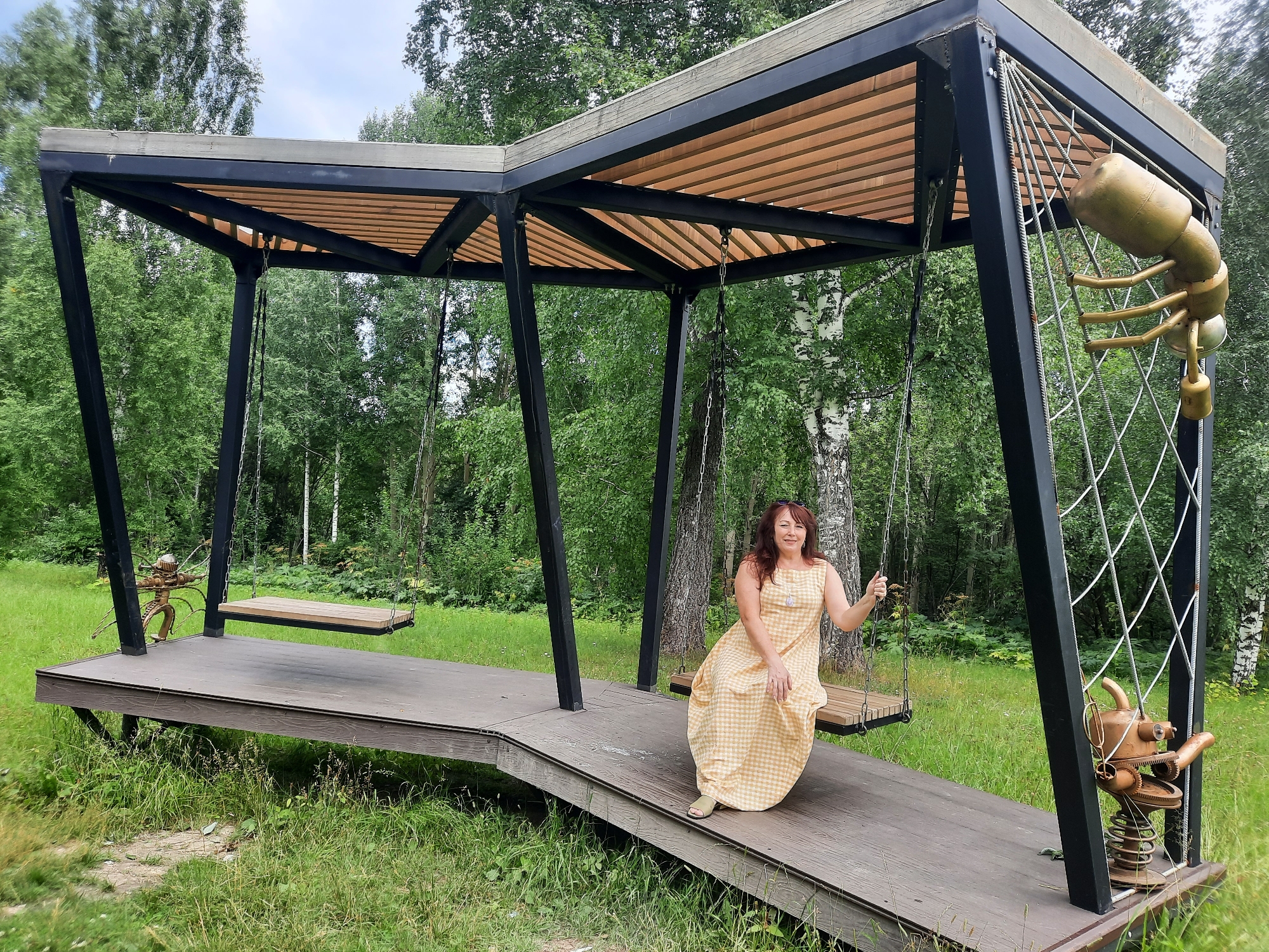 A woman with long, dark, wavy hair is sitting on a wooden swing on a raised platform. She is wearing a long, yellow, checkered dress and green sandals. The swing is suspended by metal chains from a black metal frame structure with a wooden slat roof. To the right of the swing, a large, golden, insect-like sculpture is attached to the frame. To the left, another swing is visible, as well as a smaller, metallic, insect-like sculpture. The platform is made of wood and slopes downward. The area around the platform is covered in green grass and surrounded by trees. The location is identified as Зеленогорский, Russia.