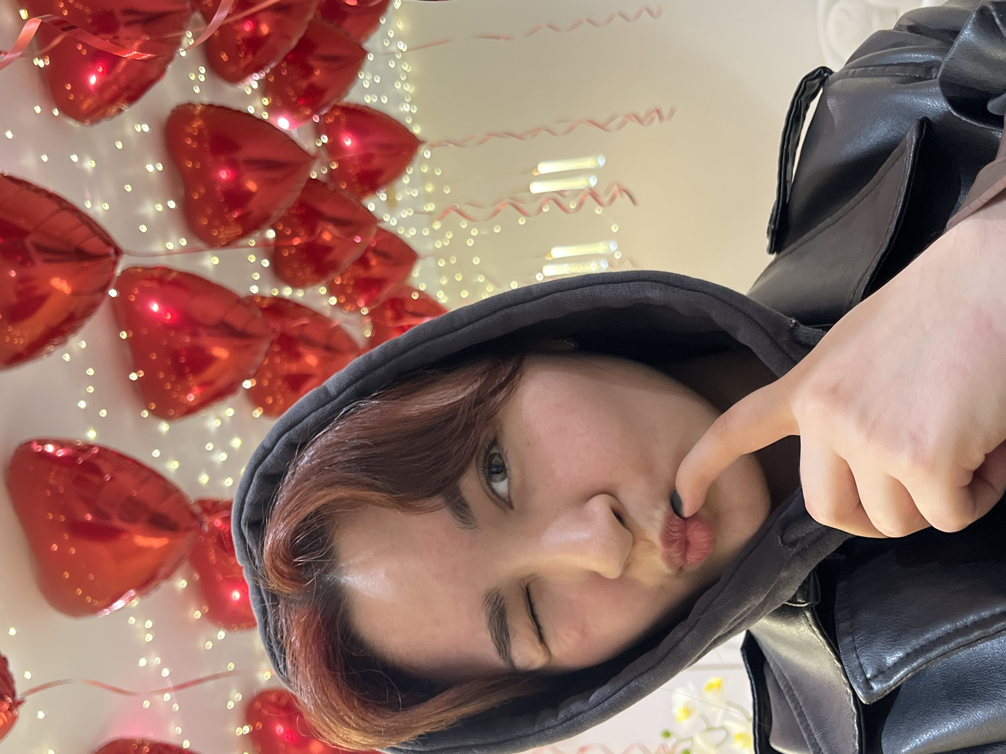 The image shows a person in a gray hoodie and a black leather jacket, winking and pursing their lips while touching them with their finger. In the background are red heart-shaped balloons and string lights. The city of Neftekamsk, Russia is not discernible in this image.