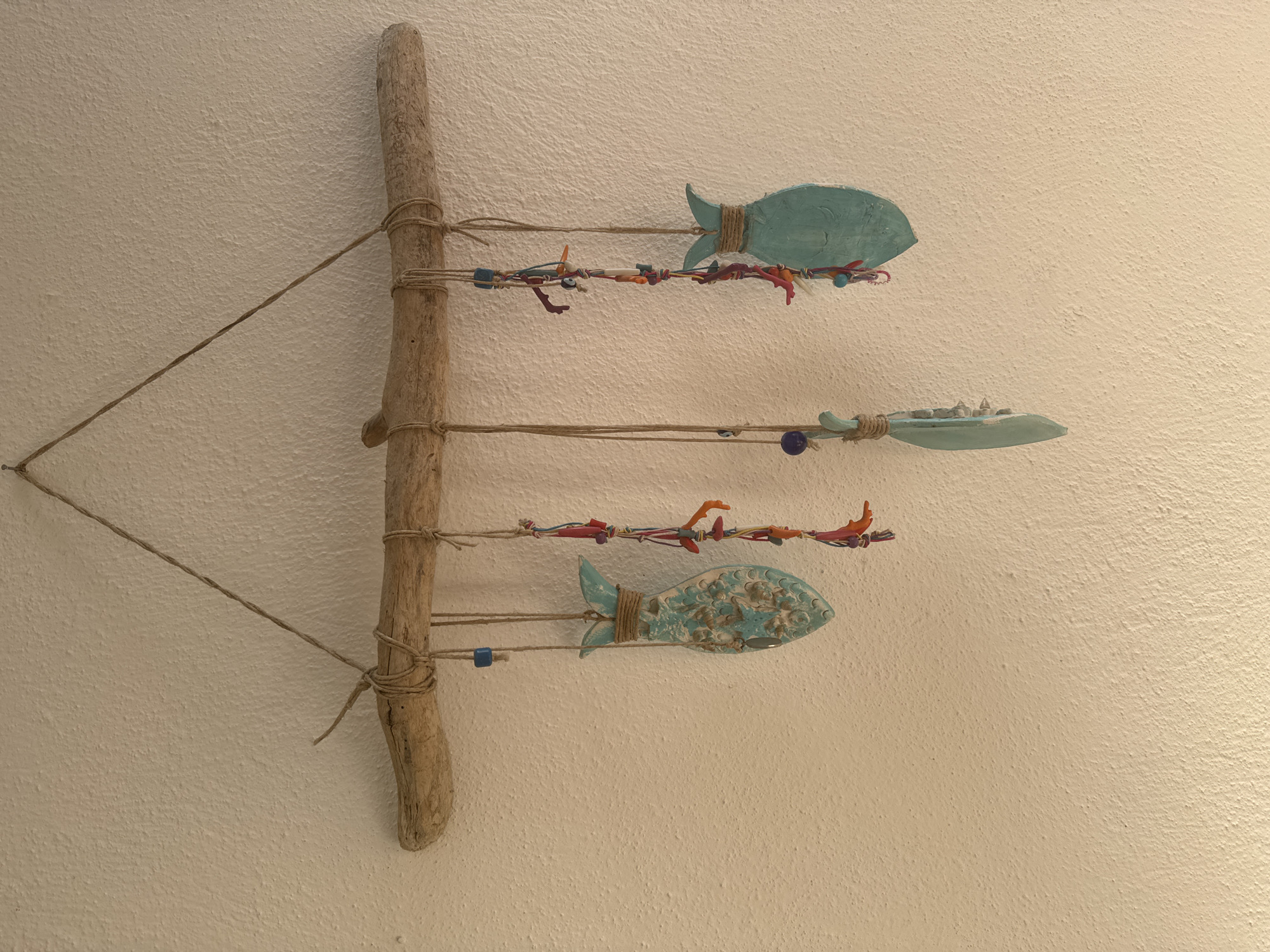 A handmade wall decoration hangs from a textured, off-white wall. The decoration consists of a piece of driftwood suspended horizontally by a piece of twine. From the driftwood, several strings of beads and ceramic fish hang. One ceramic fish is light blue with a textured pattern. Another is a solid light blue. A third ceramic element is a light blue feather shape. Other strings have colorful beads and small coral-like pieces. The location is Αμμουλιανή, Greece.