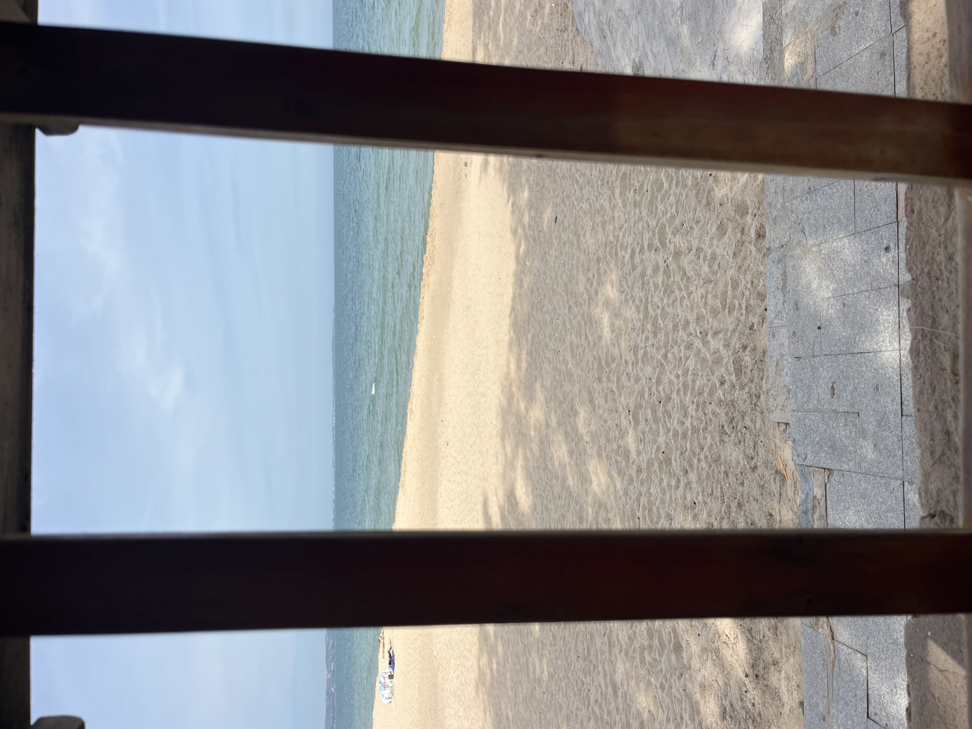 The image shows a view of a sandy beach and the sea, framed by two vertical wooden posts and a horizontal wooden beam at the top, suggesting the perspective is from a sheltered area, possibly a porch or veranda. The sandy beach is smooth with subtle wave-like textures. Further back, the sea is calm and blue, meeting a hazy horizon where land is visible. The sky is light blue with sparse white clouds. On the beach, near the water's edge, there are a few beach umbrellas and what appear to be lounge chairs, indicating a leisure area. The foreground shows a paved walkway made of grey stone slabs, some of which are weathered. The overall impression is a serene beach scene in a location that is likely a tourist resort or a private beachfront property. Based on the context provided, this scene is located in Phu Thuy Ward, Vietnam.