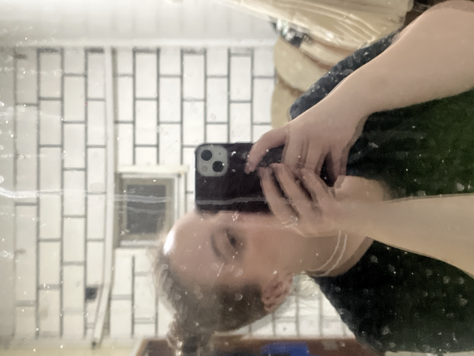 A person is taking a selfie in a bathroom mirror. The background shows white subway tiles on the walls. The location cannot be determined from the image.