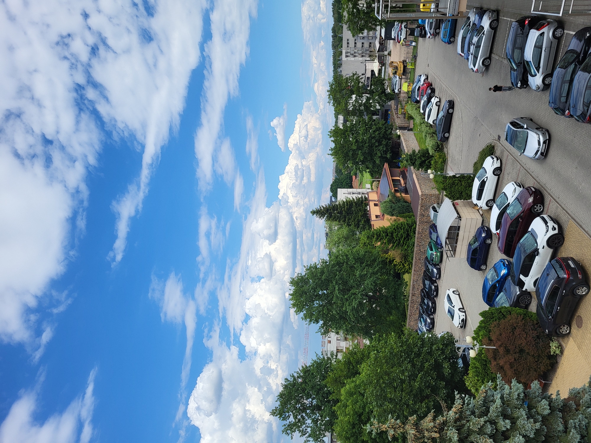 The image displays an elevated view of an urban or suburban landscape under a bright blue sky with scattered white cumulus clouds. The location is in the city of Unknown, Unknown.
The upper portion of the frame is dominated by the clear blue sky and a mix of fluffy white and some thinly stretched clouds. Below the horizon, numerous green trees, both deciduous and coniferous, form a significant part of the landscape, partially obscuring distant buildings. In the far background, behind the tree line and buildings, a red and white construction crane is visible, indicating ongoing development.
Various buildings are scattered throughout the mid-ground. These include multi-story residential or commercial structures with light-colored facades. One building in the mid-left features solar panels on its roof. A brown, textured stone wall runs horizontally across the mid-ground, separating different sections of the property.
The lower half of the image features multiple outdoor parking areas with an assortment of parked vehicles. The parking surfaces consist of both asphalt and light-colored pavers. Numerous cars of various makes, models, and colors (including white, black, silver, blue, red, and grey) are present. A white hatchback is parked prominently in the upper-middle of the paved area, and a silver hatchback is centrally located in the foreground. A yellow construction vehicle, possibly an excavator, is parked in a lot on the mid-right. One person, dressed in dark attire, is visible walking on the paved surface in the lower-middle-right section of the image, heading away from the viewer. Streetlights are present within the parking areas, and a large evergreen tree stands in the lower-left foreground, partially framing the scene.