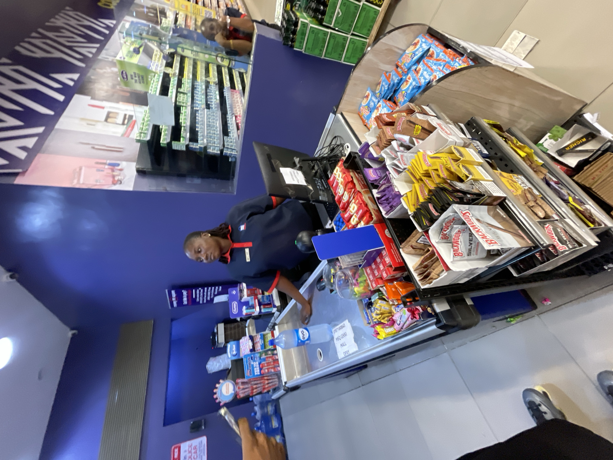 A cashier, a Black woman wearing a blue uniform shirt with a red collar and a name tag, stands behind a checkout counter. She is looking to her right. On the counter are a bottle of water, a receipt, and various products in bins. The checkout counter is in a store with blue walls and shelves stocked with products. Another person, a Black woman, is visible in the background, leaning on a counter. The scene appears to be a retail store in Moba, Nigeria.