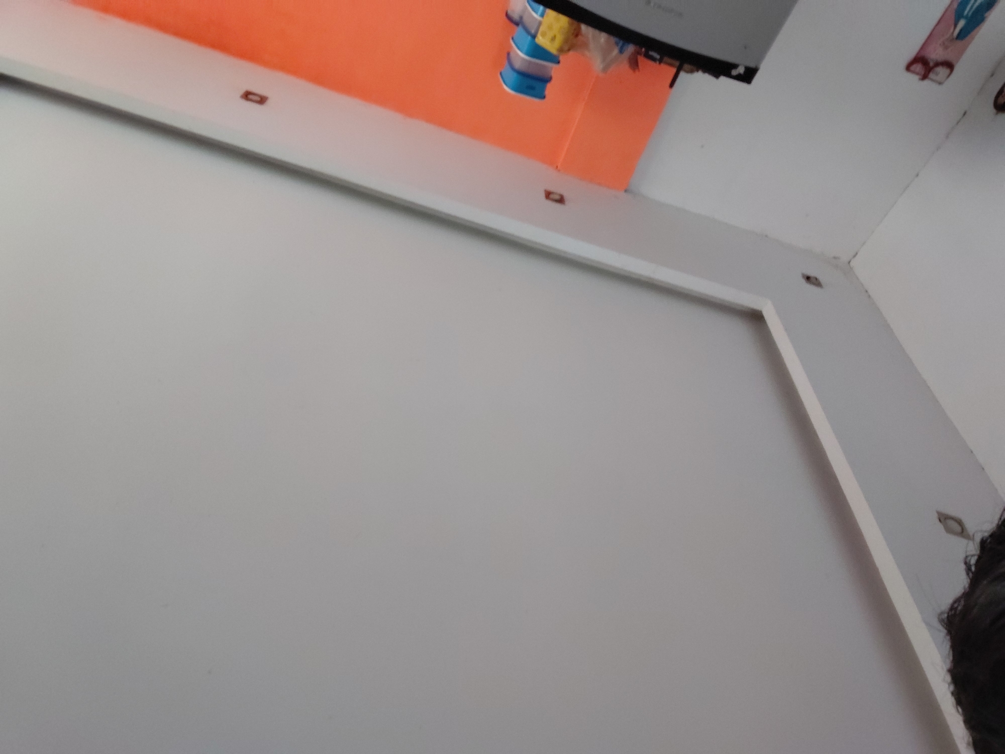 This image captures an interior view of a room in Santa Ana, Venezuela, taken from a low, upward-angled perspective. The dominant colors are white and a vibrant orange. A significant portion of the upper wall or ceiling is painted bright orange, contrasting with the surrounding white walls and ceiling.
A white, rectangular beam or architectural feature extends horizontally across the upper middle of the frame, serving as a division between white and orange surfaces. This beam contains several small, dark, rectangular inserts.
Near the top center, hanging against the orange background, are a few small, colorful objects, appearing to be plastic containers or toys in shades of blue, yellow, and white. Above these, a part of a gray or metallic appliance or fixture is visible, featuring some faint, unreadable text. To the far right, on a white wall, a small, stylized graphic or sticker depicts what looks like a figure in blue clothing with glasses, alongside a red, sandal-like item.
In the bottom right corner of the image, a section of a person's head with dark, curly hair is visible. The room appears to be evenly lit, typical of an indoor environment without direct external light sources prominent. No specific activity or event is taking place, the image shows a static view of the room's architecture and decor.