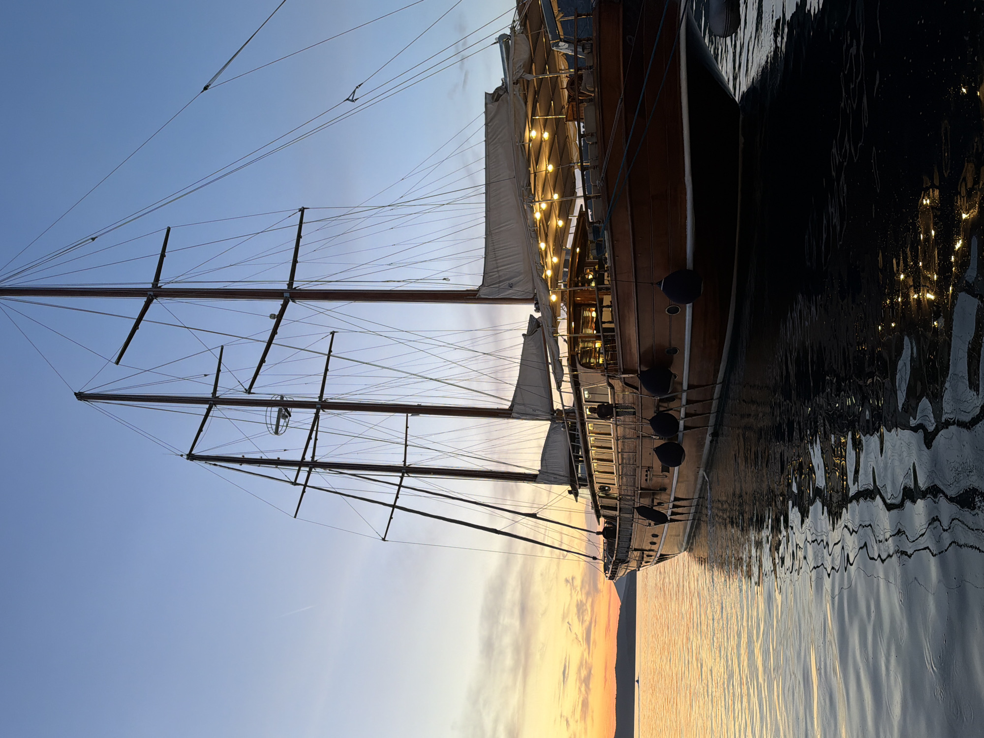 The image displays a majestic, multi-masted wooden sailing ship, likely a traditional gulet, at rest in what appears to be a harbor or bay in Poros, Greece. The photograph is taken at either sunrise or sunset, evidenced by the vibrant colors of the sky and the atmospheric lighting.
The sky transitions from a clear, light blue directly overhead to warm, soft hues of orange, yellow, and pink near the horizon, suggesting the sun is low. A distant landmass, possibly hills or mountains, is silhouetted against this colorful backdrop.
The ship, the primary subject, features a dark, polished wooden hull and prominent masts with an intricate web of rigging and spars. White sails are visible, either furled or partially furled. A notable detail is the string of warm-toned lights illuminating the ship's deck area, casting a inviting glow. These lights, along with the sky's colors, are beautifully reflected in the calm water surface below. Several dark fenders hang along the side of the ship's hull, visible above the waterline.
The water itself is tranquil, displaying gentle ripples that shimmer with the reflections of the sky and the ship's lights. The darker sections of the water vividly reflect the ship's hull. No people are visible, but the illuminated deck lights imply a readiness for evening activity or recent human presence. There is no visible text in the image. The overall scene conveys a sense of peaceful beauty and the charm of maritime life during the golden hour.