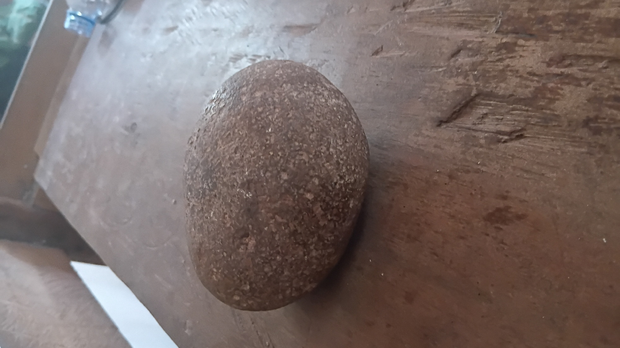 A brown, oval-shaped stone rests on a rustic wooden surface. The stone exhibits a textured, slightly rough exterior. The wooden surface appears weathered, with visible grain and some markings. In the background, out of focus, elements of a room are discernible, including what might be furniture or walls, and a hint of greenery through a window, suggesting an indoor setting. The lighting suggests it is daytime. No individuals or specific actions are depicted. The scene is static and focused on the stone and its immediate surroundings. The image is taken from a slightly elevated angle. It is not possible to determine the exact location within Prabumulih, Indonesia based on the visual information provided.