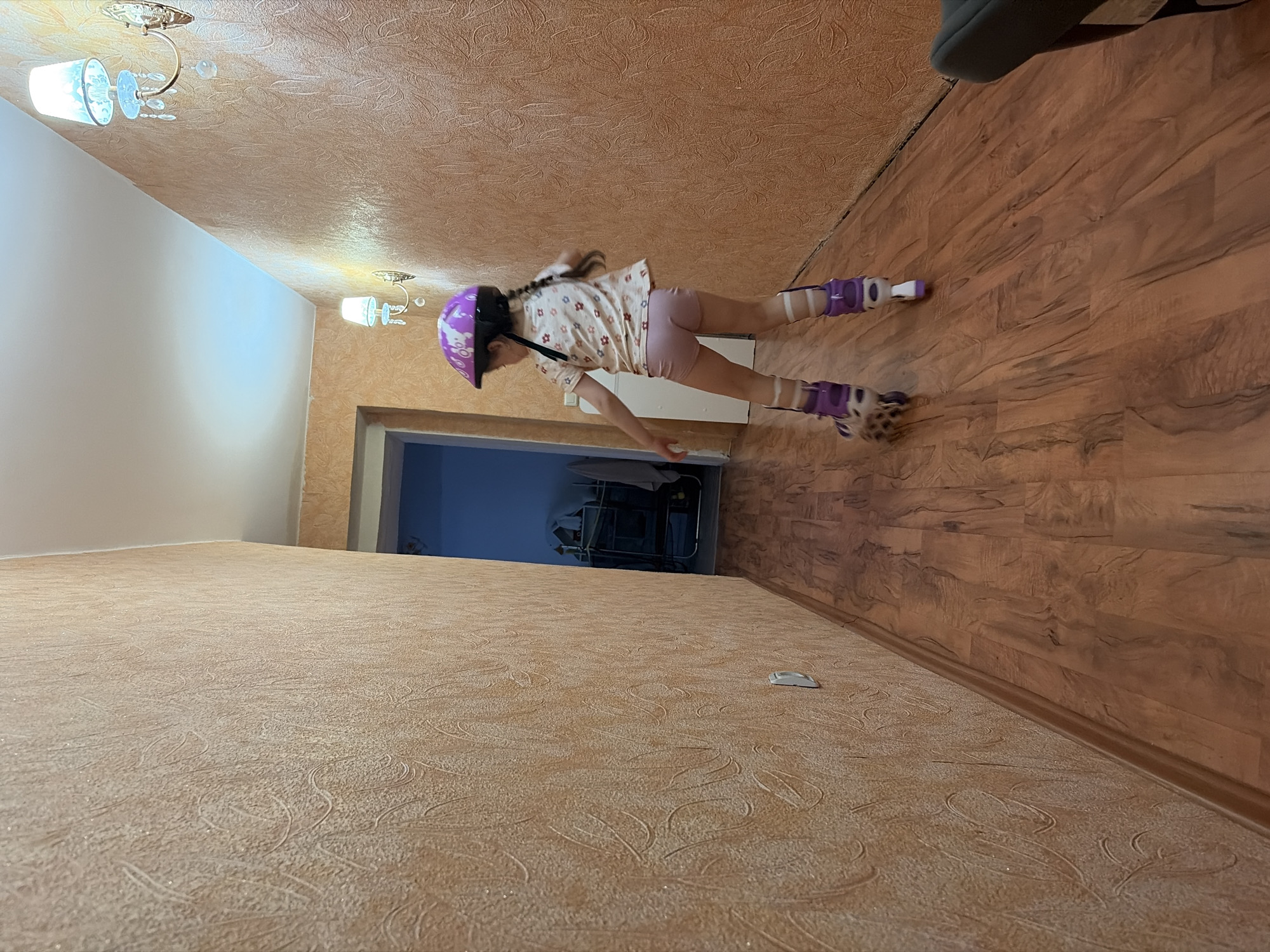 A girl wearing a purple helmet and inline skates is inline skating from an open doorway onto a wooden floor. The room has a light-colored textured wall and a textured ceiling. The open doorway leads to another room with blue walls and a rack. There is a light fixture on the ceiling. The photo was taken indoors. The photo appears to have been taken in a residential space, likely in Chaikovsky, Russia.