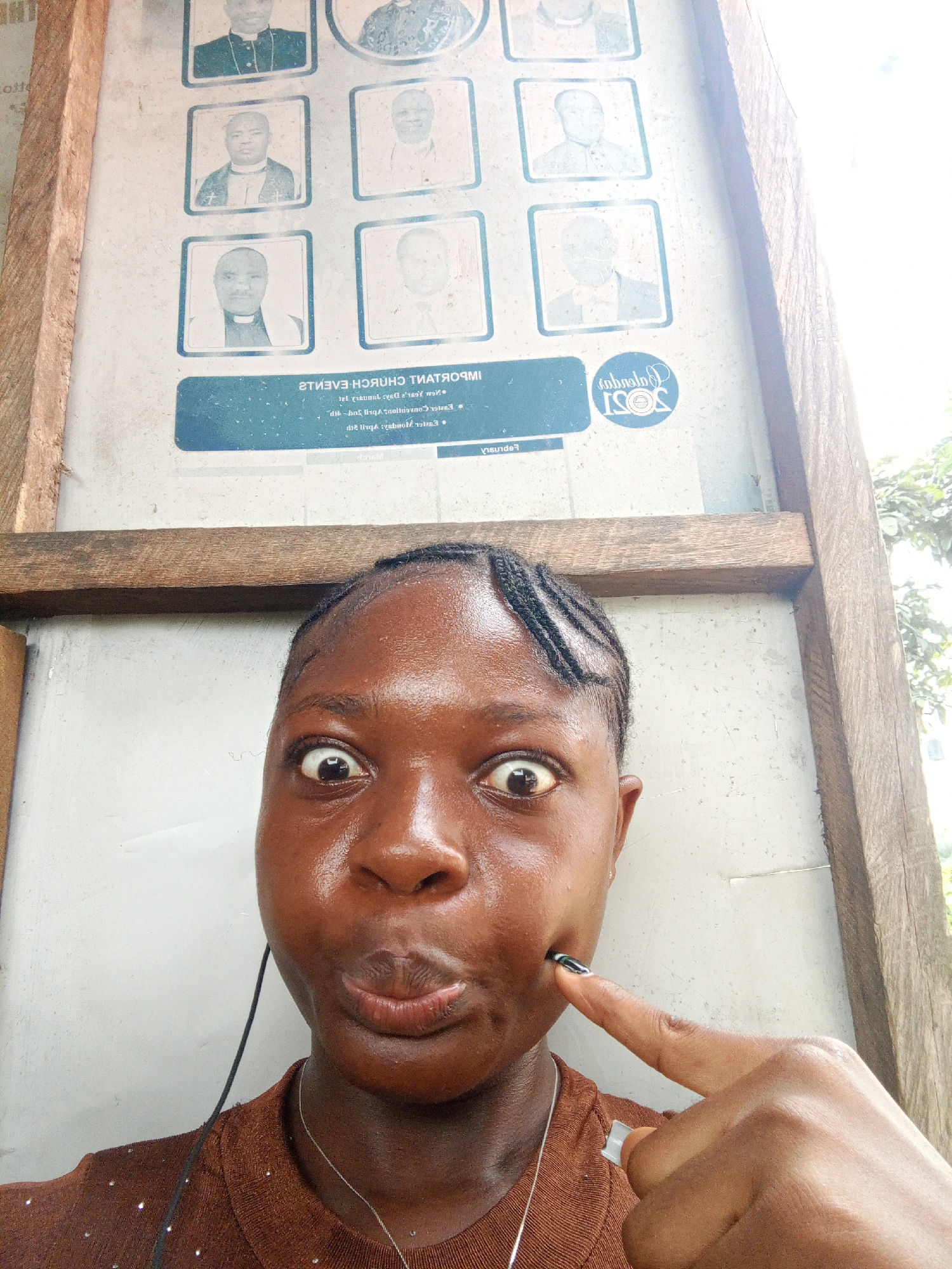 A dark-skinned young woman is the subject of a close-up, selfie-style photograph taken outdoors, in what appears to be Mbribit, Nigeria based on the text in the upper portion of the photo. She is positioned in front of a weathered bulletin board, possibly belonging to a church, with a wooden frame. The board displays several portrait photographs of men in clergy attire. In the lower portion of the board, reversed text indicates important church events, with the word "IMPORTANT" being visible in reverse. Below this is a logo for "ISOS Gabrain." The woman has braided hair and is wearing a brown, possibly glittery, top with a silver necklace. Her skin is slightly shiny, and she is making a playful face with wide eyes and pursed lips, while pointing to her right cheek with her index finger, which has a dark polished nail. Her expression and gesture suggest a lighthearted and humorous mood. A thin cord, possibly from headphones, is visible draped down the front of her shirt. The lighting appears bright, suggesting daytime. Part of a tree is slightly visible.