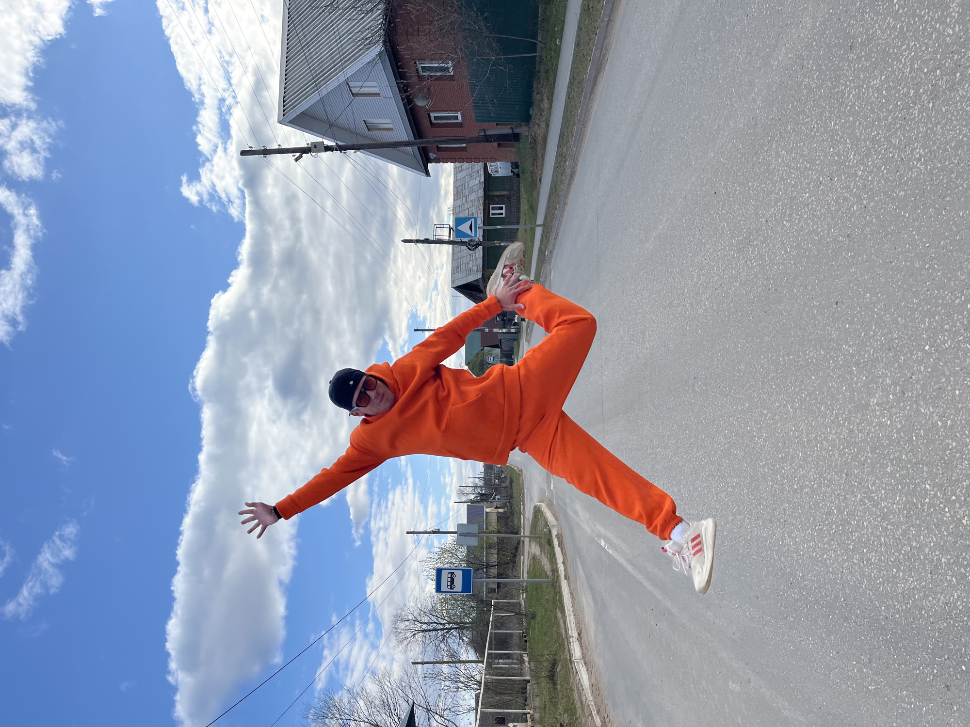 A man in an orange tracksuit, white sneakers, and sunglasses balances on one leg in the middle of a road in what appears to be a rural area near Chaikovsky, Russia. The man has one arm outstretched towards the bright blue sky with cumulus clouds. The other leg is bent up and the foot is being held by the man's hand. In the background, there is an asphalt road, houses, telephone poles, and a bus stop with a blue sign.