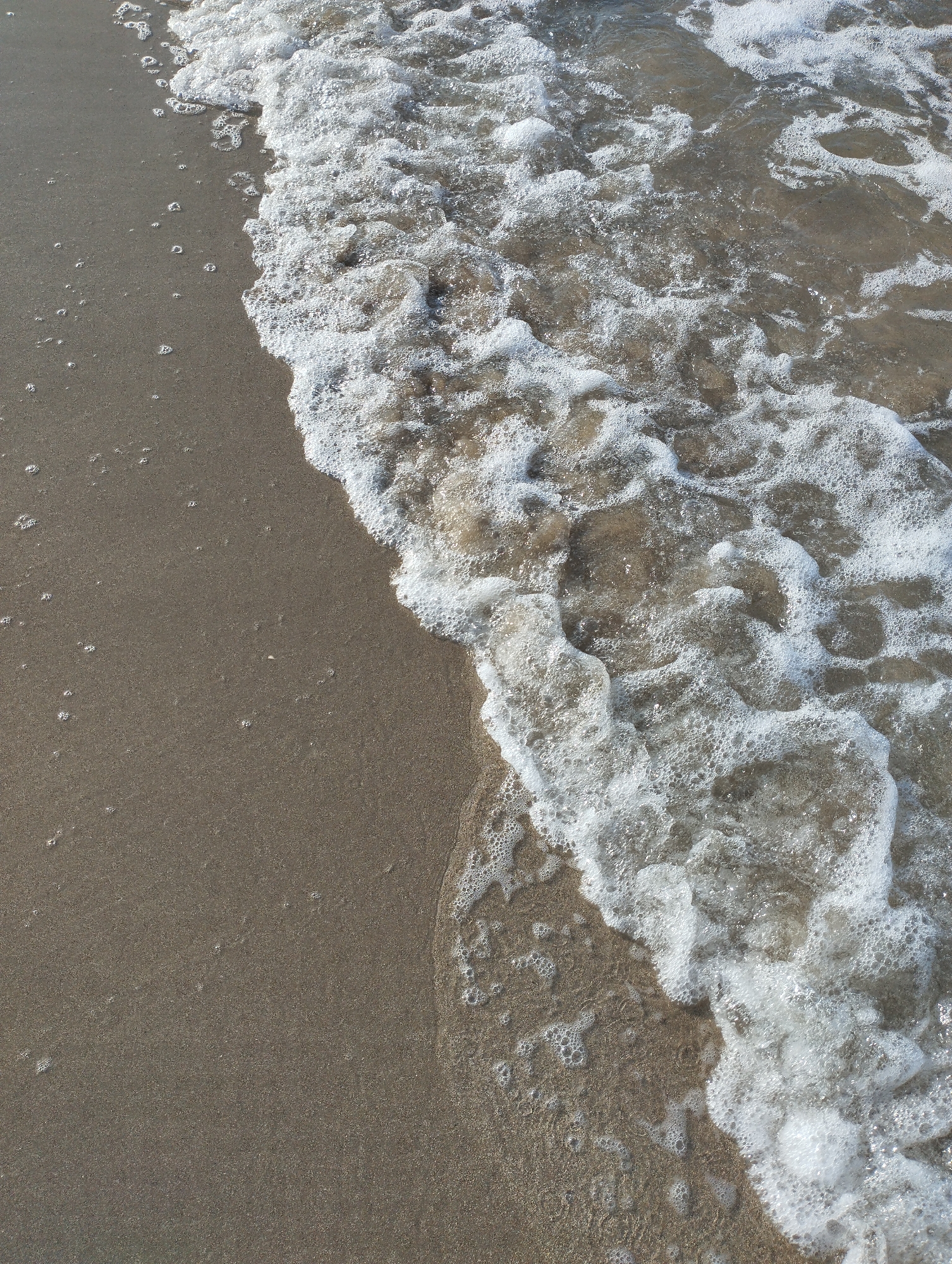 An extreme close-up, high-angle shot captures the edge of the ocean meeting a sandy beach. The water, appearing shallow, is filled with white foam and bubbles, indicating the presence of waves breaking or receding. The sand is a muted brown color with a slightly wet appearance where it meets the water. Small bubbles are also scattered across the wet sand. The overall scene is one of natural coastal movement, with no people or man-made objects visible.