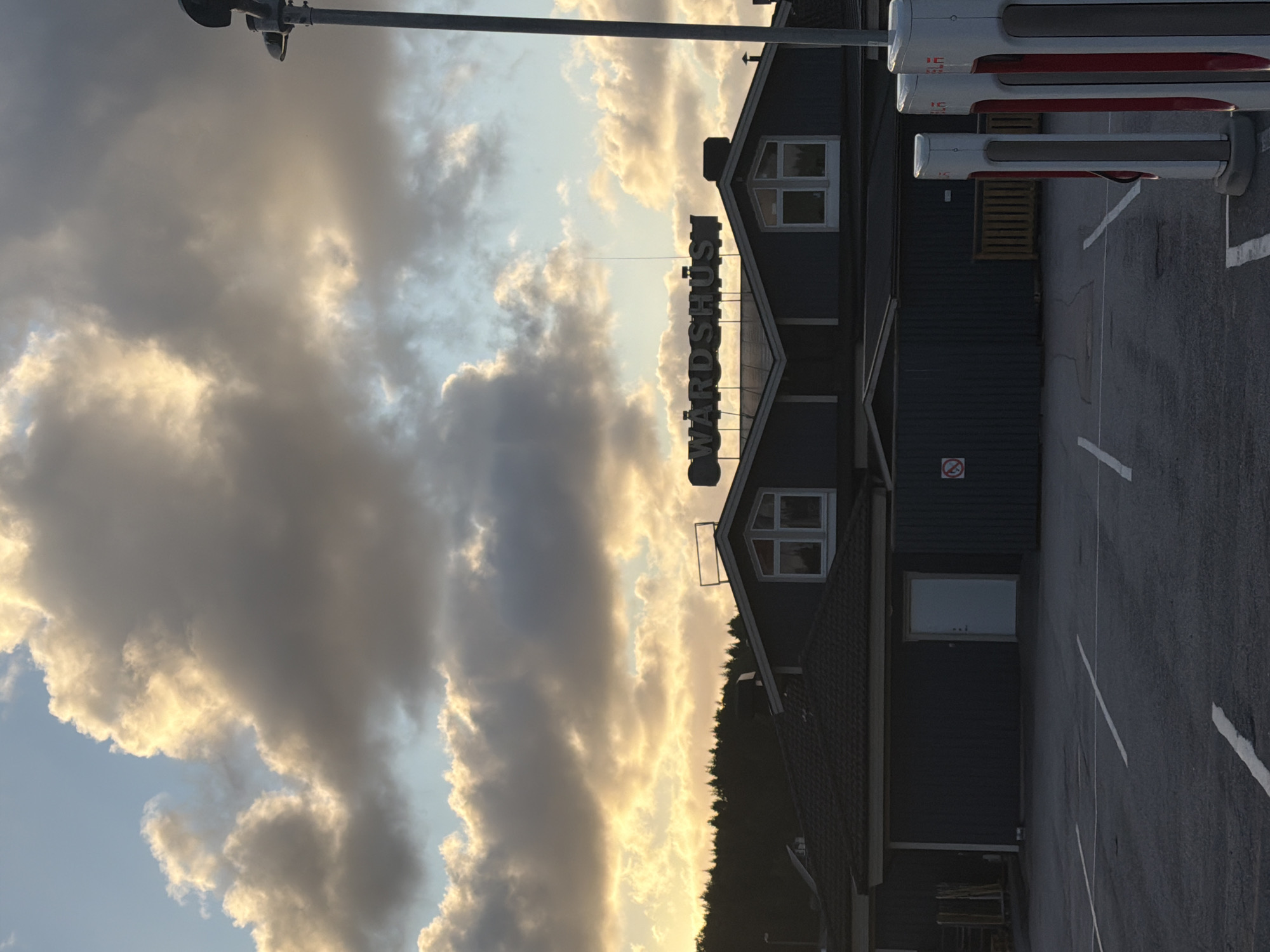 In the city of Hogstorp, Sweden, a parking lot is pictured with several Tesla charging stations in the foreground. In the background, a dark-colored building with a prominent sign reading "VÄRDSHUS" is visible, situated against a backdrop of dark clouds illuminated by the setting sun. A line of trees forms a silhouette behind the building, and a lamppost stands to the right of the frame. The parking lot is marked with white lines.
