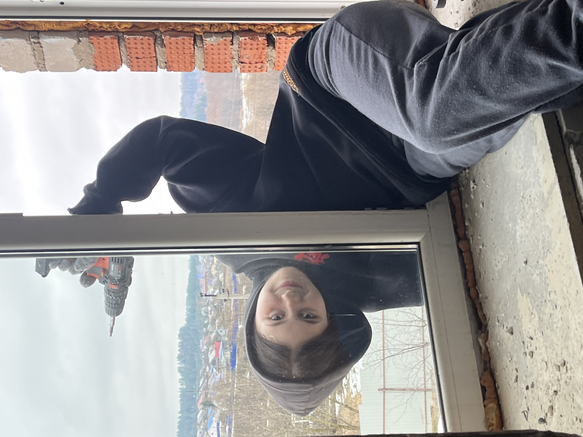 A person is installing a window. The person in the window reflection is facing the camera. A drill is being held up outside the window and is reflected in the window's glass. Outside the window is a neighborhood in Фоки, Russia. The brick wall can be seen above the window, with brick-shaped insulation.