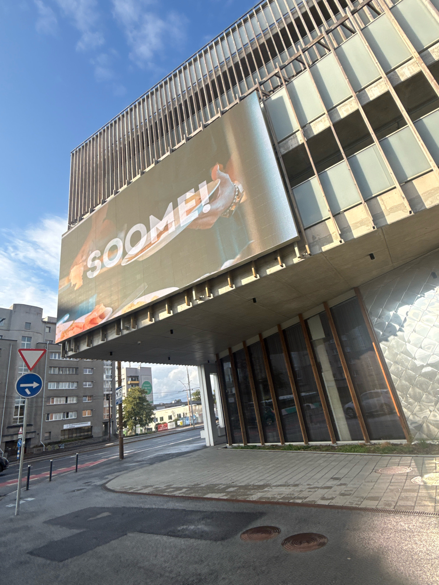 The image captures a street-level view of a modern building with a large digital billboard displaying the word "SOOME!" in bold white lettering. The billboard is positioned high on the building, overlooking a wet asphalt road and sidewalk. The building itself features a contemporary architectural style with a facade of glass panels and metal framing, suggesting it is a commercial or public structure.
In the background, several other buildings are visible, typical of an urban environment. To the left of the main building, a traffic sign with a red triangle and a blue circle with a white arrow pointing right is mounted on a pole. This indicates traffic regulations for the area. The sky is partly cloudy, with patches of blue visible, suggesting daytime.
The scene appears to be a section of the city with moderate urban development, characterized by multi-story buildings, paved roads, and sidewalks. The wet surfaces of the road and sidewalk indicate recent rain or cleaning. There are no people or vehicles prominently featured in the immediate foreground or on the billboard, focusing the attention on the architectural elements and the advertisement. The overall impression is a snapshot of a dynamic urban landscape in Tallinn, Estonia, highlighted by modern architecture and commercial display.