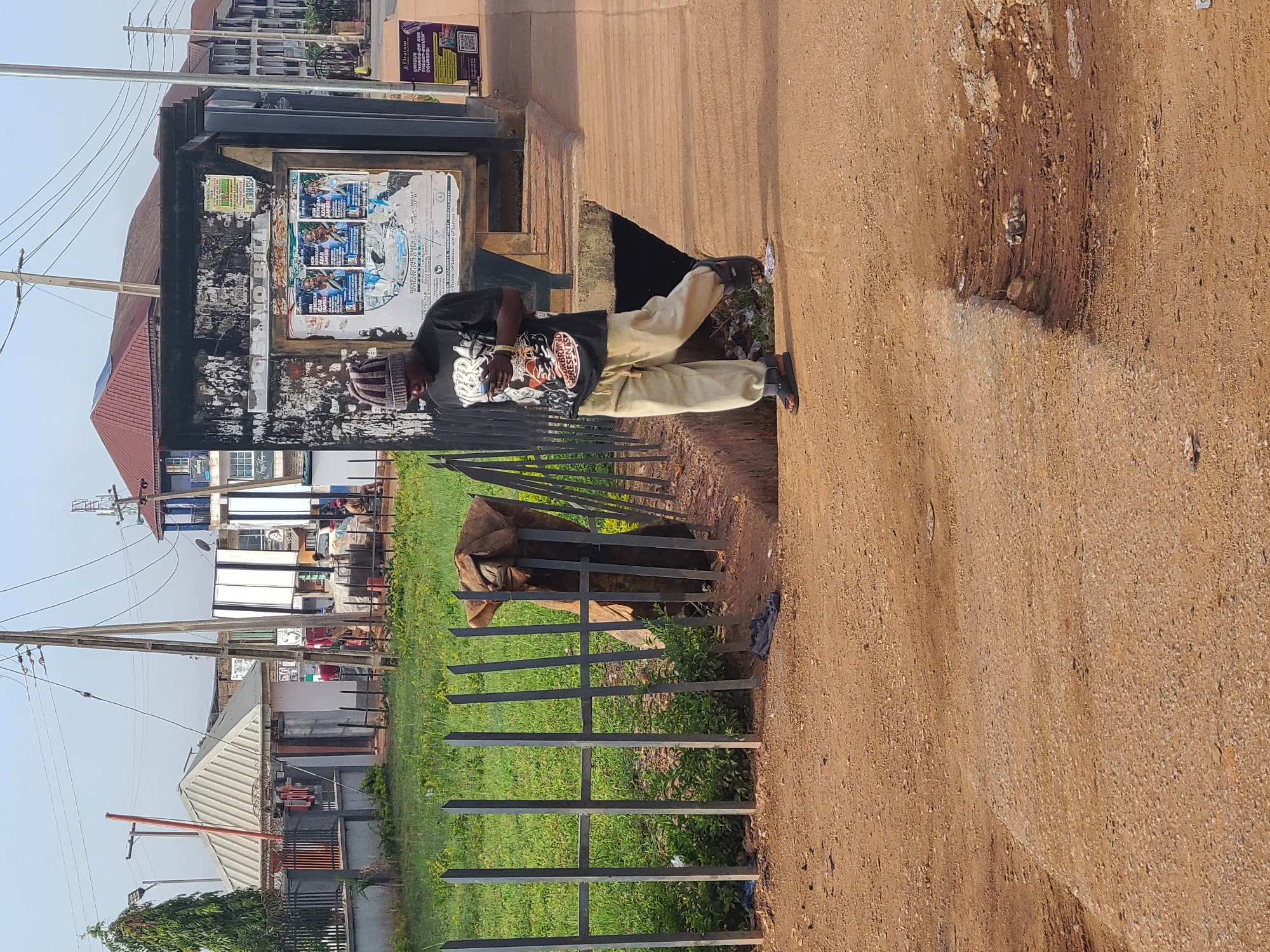 The image shows a man standing near a metal fence on the side of a dirt road in what is likely Gura Top, Nigeria. The man is wearing a black t-shirt, khaki pants, and sandals, and appears to be looking at something in his hands. Behind the fence, there is a grassy area and what looks like the head of a brown cow poking through the bars. In the background, there are buildings and utility poles. To the left of the man, there is a bus stop with posters attached to it. The road surface is dirt, with a significant drop-off at the edge.
