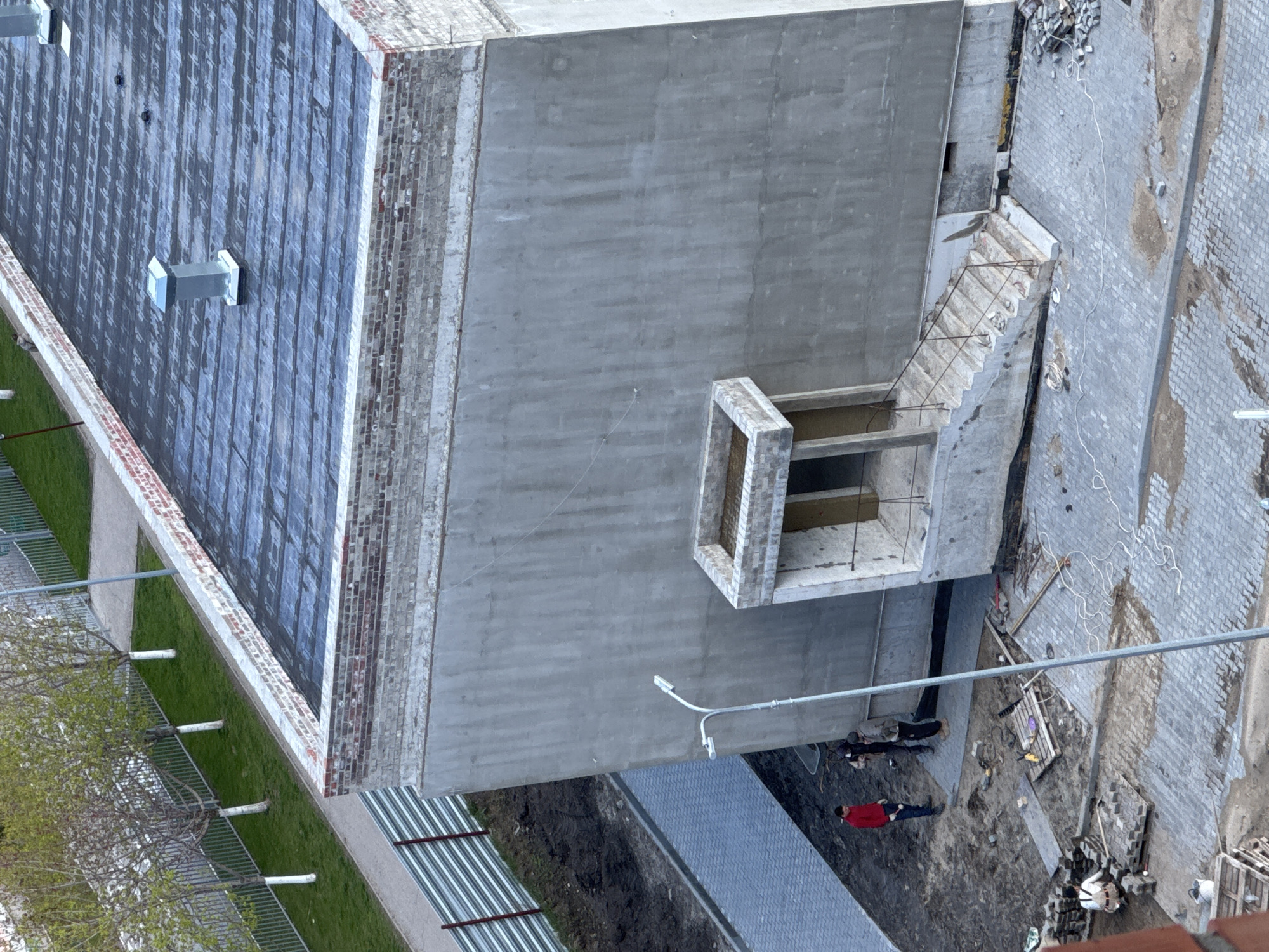 The image shows an aerial view of a construction site in an urban environment. The location is in a city named Unknown in Unknown.
The scene is dominated by a concrete building under construction. To the left, there is a roof with visible ventilation units and a brick facade. A set of unfinished stairs with rebar is attached to the building. Below the building, corrugated metal panels are visible, presumably for construction purposes.
On the ground, several people are present. One person is wearing a red shirt. Construction materials, debris, and construction tools are scattered around the work site. The ground is a mix of dirt and paved areas. Overhead power lines are visible in the lower center of the image.