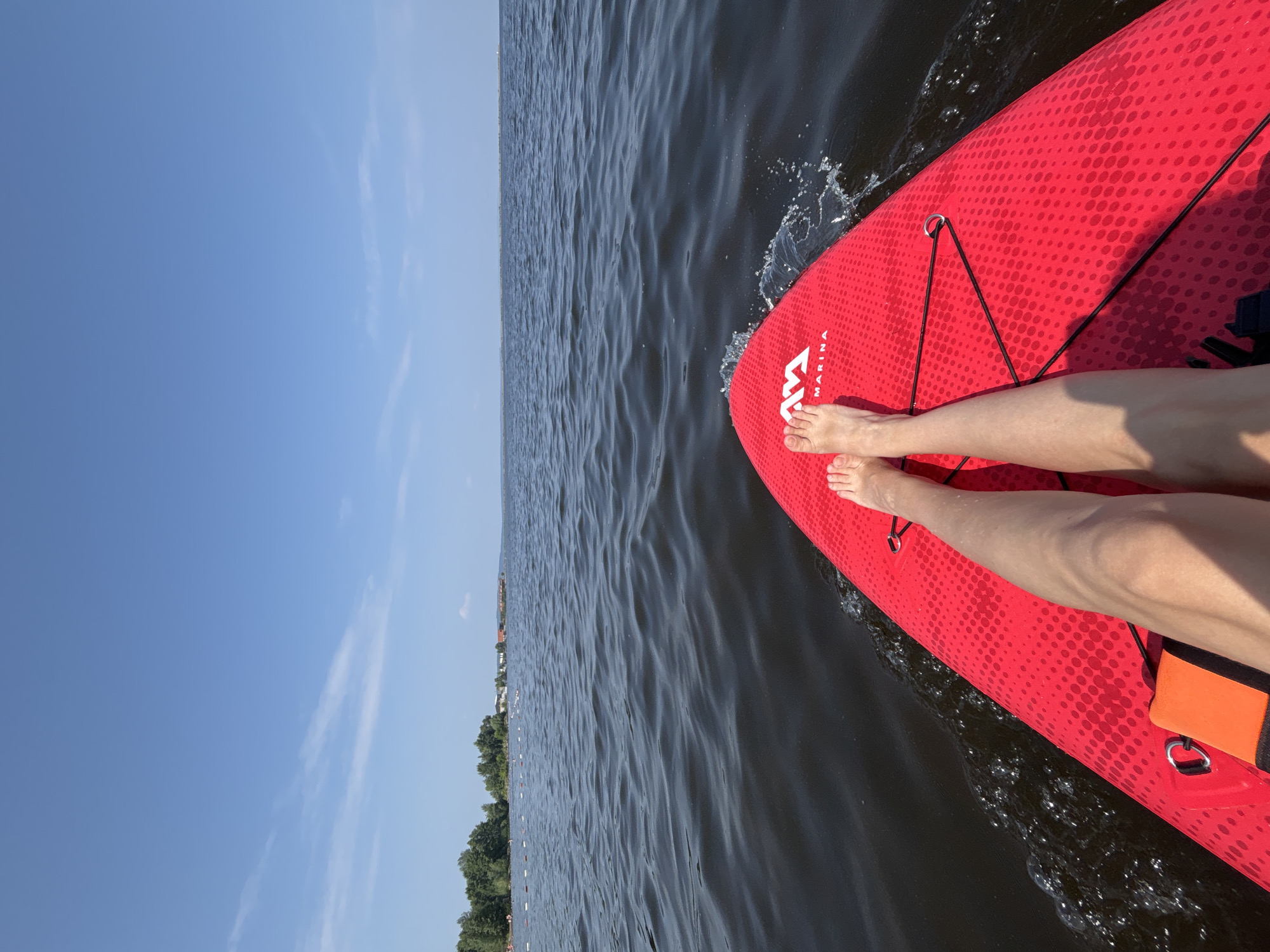 The image shows a person's legs and feet on a red paddleboard in dark water. The paddleboard has a dotted pattern and a logo that reads "M MARINA." The water is rippling, and some splashes are visible. In the background, a line of trees and buildings can be seen along the shore under a clear blue sky with a few wispy clouds. The scene suggests a sunny day, likely in the afternoon, with calm weather suitable for water activities. The person appears to be enjoying a paddleboarding excursion, with a relaxed and peaceful atmosphere. The overall impression is one of a serene summer day spent on the water.