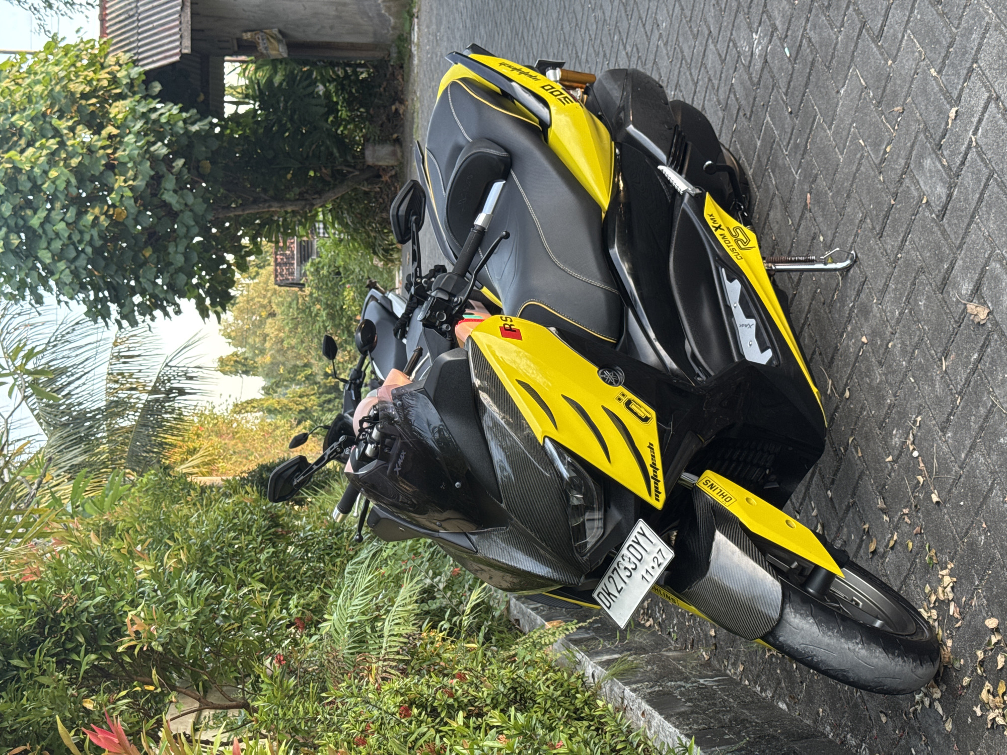 The image shows a Yamaha TMax scooter parked on a brick or stone paved road next to lush green vegetation. The scooter is predominantly yellow and black, featuring visible decals and a license plate that reads "DK2733DYY 11.27". The scooter's tires are black and appear to be slightly worn.
The background suggests an outdoor environment, with greenery extending beyond the scooter and a partial view of a building structure. Based on the license plate format and the visible vegetation, the location is consistent with Kuta Selatan, Indonesia. There are no visible people in the frame. The scooter is the main subject. The only activity implied is the scooter being parked.