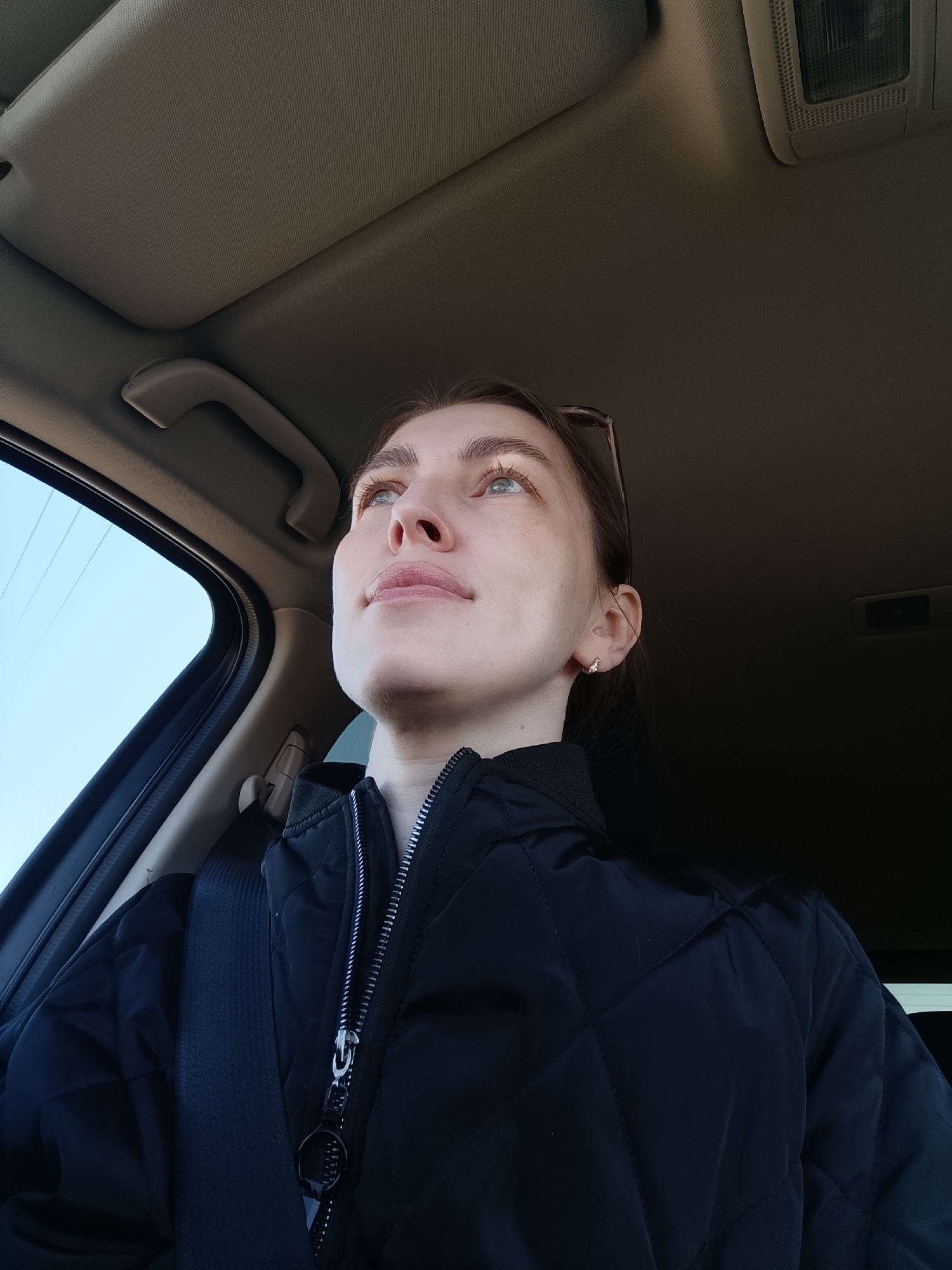 A young woman is seated in a car, wearing a black quilted jacket and a seatbelt. She is looking upwards and towards the left. The car interior is visible, including the roof and window. Outside the window, a clear blue sky is visible. There is no information to indicate the location is Chaikovsky, Russia.
