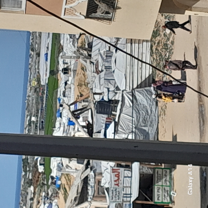 The image provides an elevated view of a dense, informal settlement within the city of Khan Yunis, Palestinian Territory, extending towards the Mediterranean Sea visible on the horizon.
In the immediate foreground, a dark gray vertical pillar partially obstructs the left-center view. To the right, a portion of a light-colored, multi-story building is visible, featuring a window with a metal grate and ornate white decorative elements. A dark cable extends diagonally across the frame from the upper right.
The midground features an unpaved ground area where five individuals are present. A person in a dark purple abaya and head covering stands facing right, accompanied by another individual in lighter attire and head covering, and two children, one in a yellow top. A male figure in a dark jacket and pants walks towards the right. Another male figure in a light brown jacket and dark pants walks towards the left.
To the left of the individuals, a makeshift stall or shop structure is visible with a white awning and a sign in Arabic displaying "المجد" (Al-Majd). Below it, a green and white sign shows "PAY PAL" and "SDF" along with other text.
Behind these foreground elements, a vast expanse of densely packed, informal shelters and tents dominates the scene. These structures are predominantly constructed from white and light-colored tarpaulins stretched over frames, interspersed with wood, corrugated materials, and exposed sandy ground. Black water tanks are visible on the roofs of some shelters.
In the background, the informal settlement continues, giving way to green agricultural fields and large white-covered structures, likely greenhouses. The furthest visible element is a clear blue sky meeting a distinct line of a darker blue body of water, identifying it as the Mediterranean Sea. The scene is brightly lit by natural daylight.
