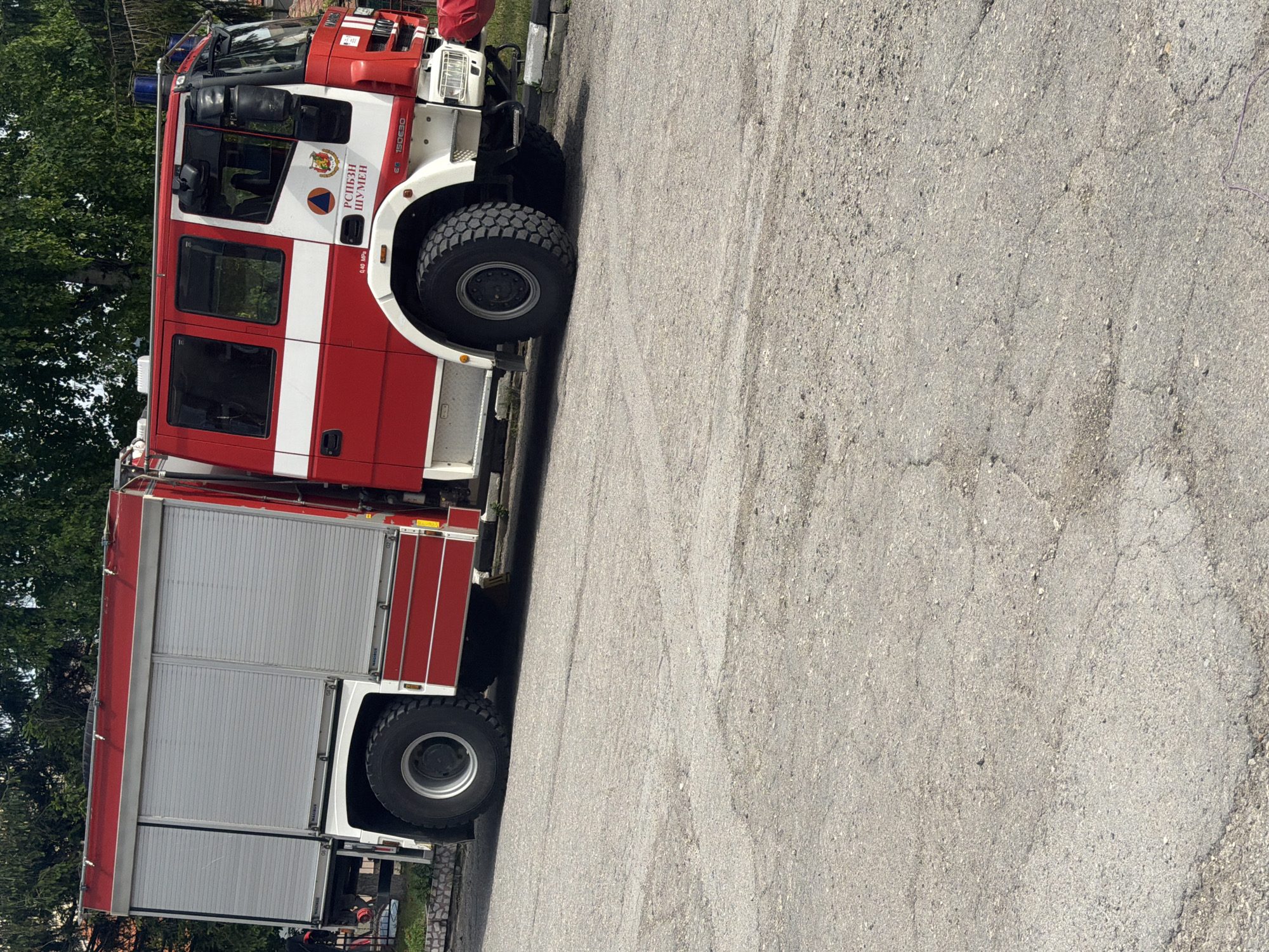 The image displays a large, red and white fire truck parked outdoors on a grey asphalt surface, likely in Shumen, Bulgaria. The truck is prominently featured, showing its full length from the front cab to the rear. It has a distinctive red and white livery with a white stripe running along its side. The cabin is red, and the truck is equipped with large, heavily-treaded tires suitable for various terrains. Blue emergency lights are visible on the roof of the cab.
The main body of the fire truck is red with multiple silver-grey roll-up compartment doors along its side, some of which bear the faint "MAGIRUS" branding, suggesting the manufacturer.
Visible text on the side of the truck's cabin, written in Cyrillic, clearly states "ГД ПБЗН" (General Directorate Fire Safety and Civil Protection) and "ШУМЕН" (Shumen), confirming its operational location in Shumen, Bulgaria. An official emblem featuring a shield and a crest, along with a triangle within a circle symbol, is also present near the text. Additional markings include "150E30," which is likely the vehicle model number, and "0.40 MPa," possibly indicating a pressure rating.
The fire truck appears stationary, parked on the asphalt. No people are visible, and no specific activity or event is taking place. The background shows green foliage from trees, indicating an outdoor setting. The lighting suggests it is daytime with clear weather. The asphalt surface exhibits some signs of wear and cracks.