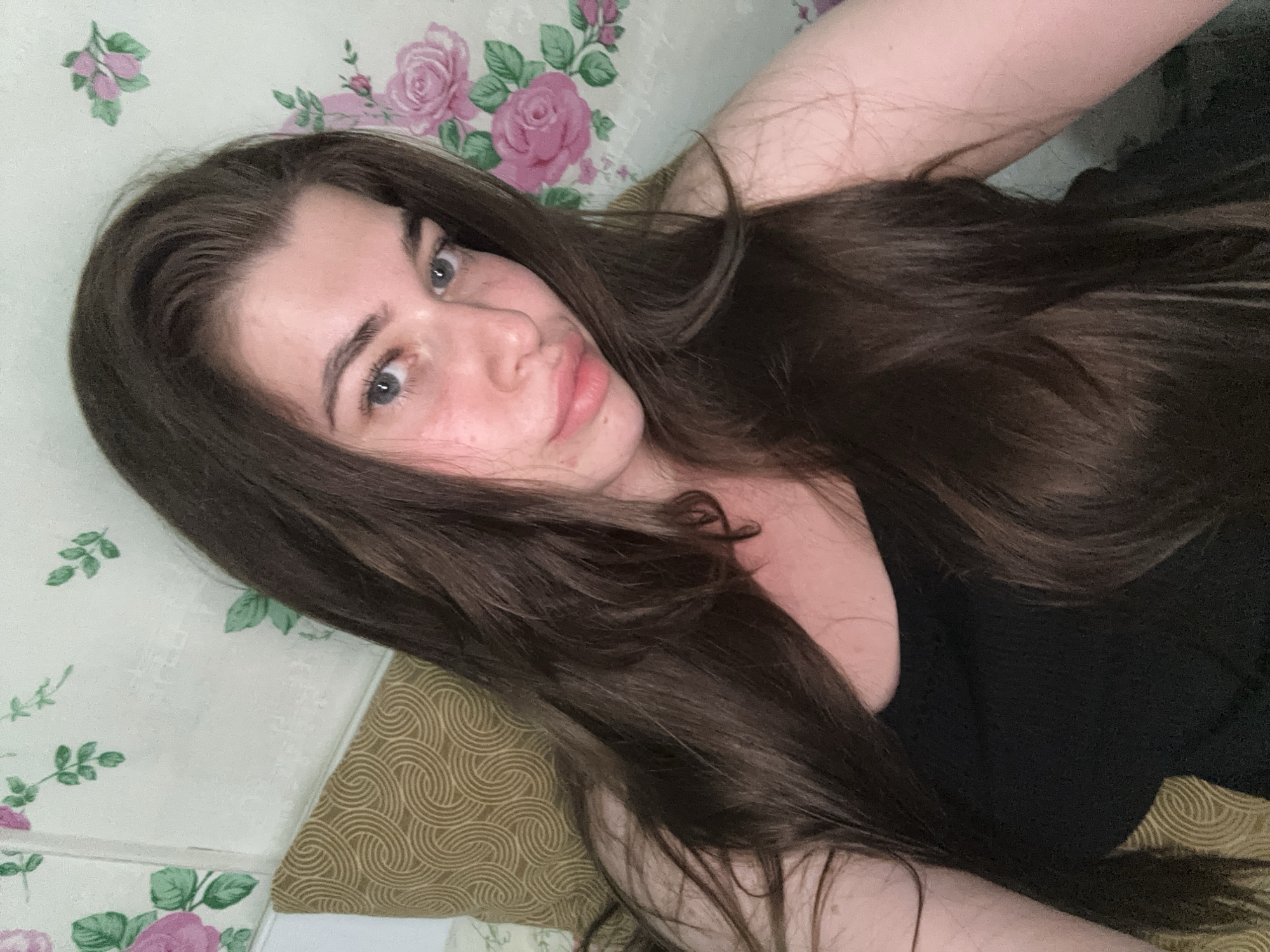 A young woman with long, dark brown hair and blue eyes is shown in a close-up, selfie-style portrait. She is wearing a black tank top. Her skin appears flushed, and she has some freckles. Behind her is a wall with a floral wallpaper featuring pink roses and green leaves. A textured, gold-colored pillow is partially visible on her left. The image does not contain information that allows for the specific location in the city of Мещёры, Russia to be determined.