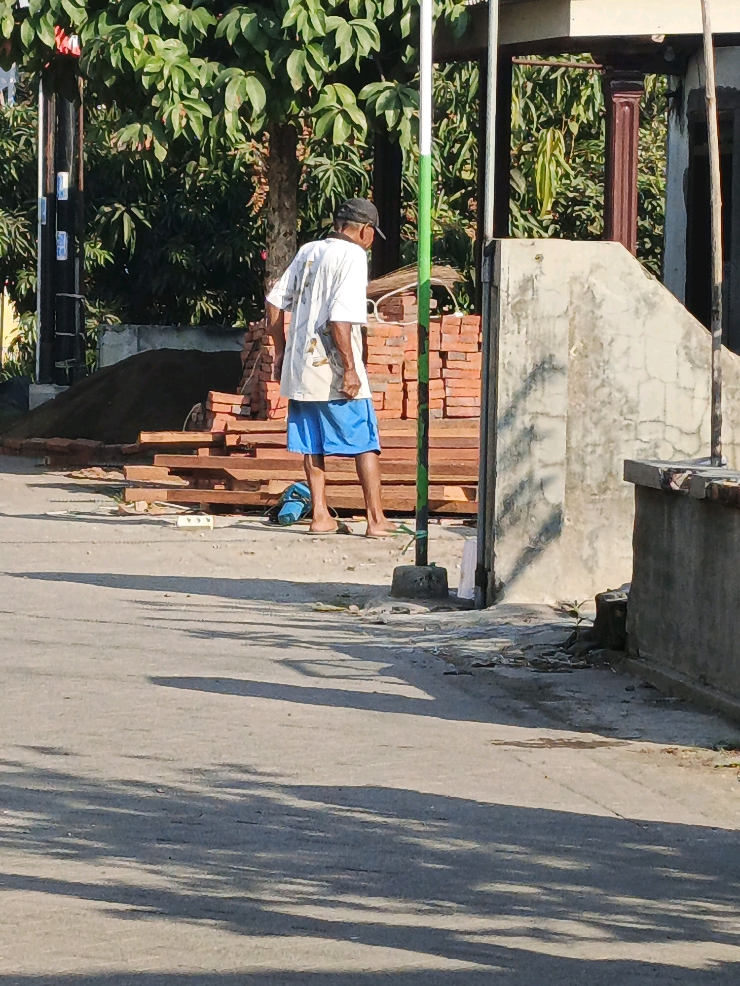 In Ketegan, Indonesia, an adult male, wearing a white t-shirt and blue shorts, stands facing away from the camera on a paved road. He is barefoot and appears to be holding a broom or similar cleaning tool. To his right, a pile of red bricks and wooden planks are stacked near a partially constructed concrete structure. Behind the man and the construction materials, lush green foliage and trees are visible, suggesting an outdoor, possibly residential or developing area. The scene is illuminated by bright sunlight, casting long shadows across the road. A blue bag is on the ground near the man.